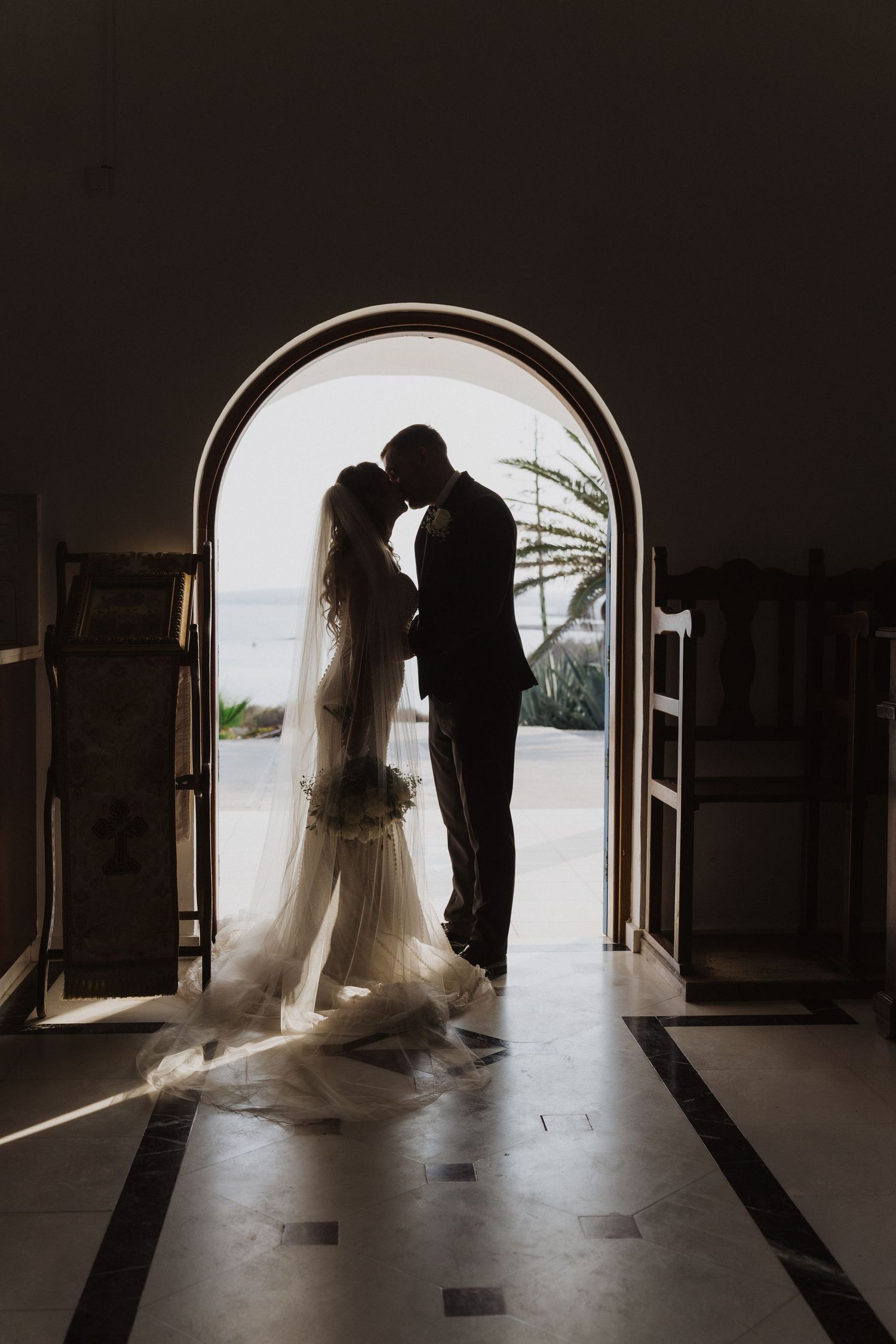 Silhouette of bride and groom embracing in an arched doorway with ocean light behind them.
