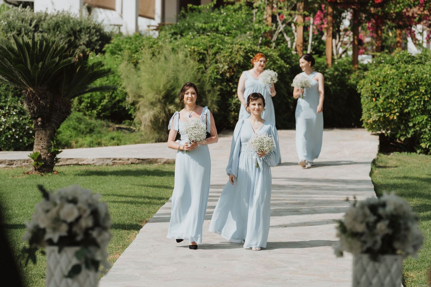 Bridesmaids in light blue gowns walking down pathway carrying white bouquets.