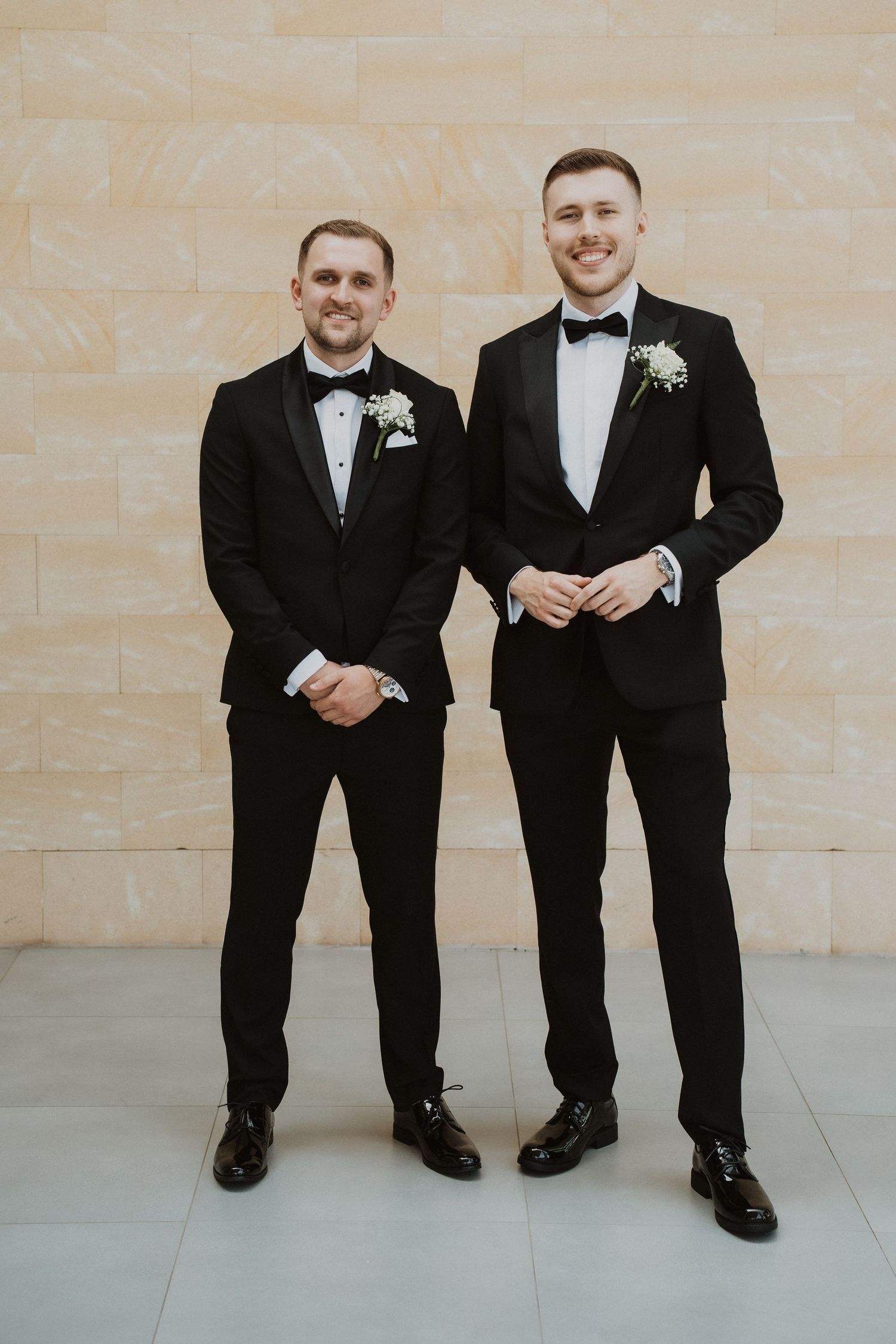 Two groomsmen in matching black tuxedos with white boutonnieres posing together.