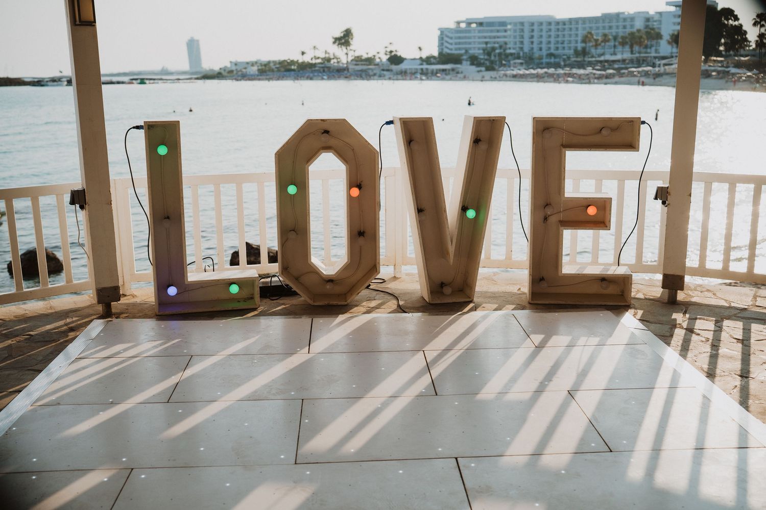 Large illuminated LOVE marquee letters on a dance floor with waterfront and palm trees in the background.