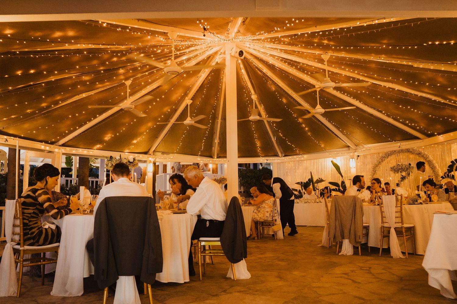Guests mingle under a warmly lit transparent tent with fairy lights at an elegant evening wedding reception.
