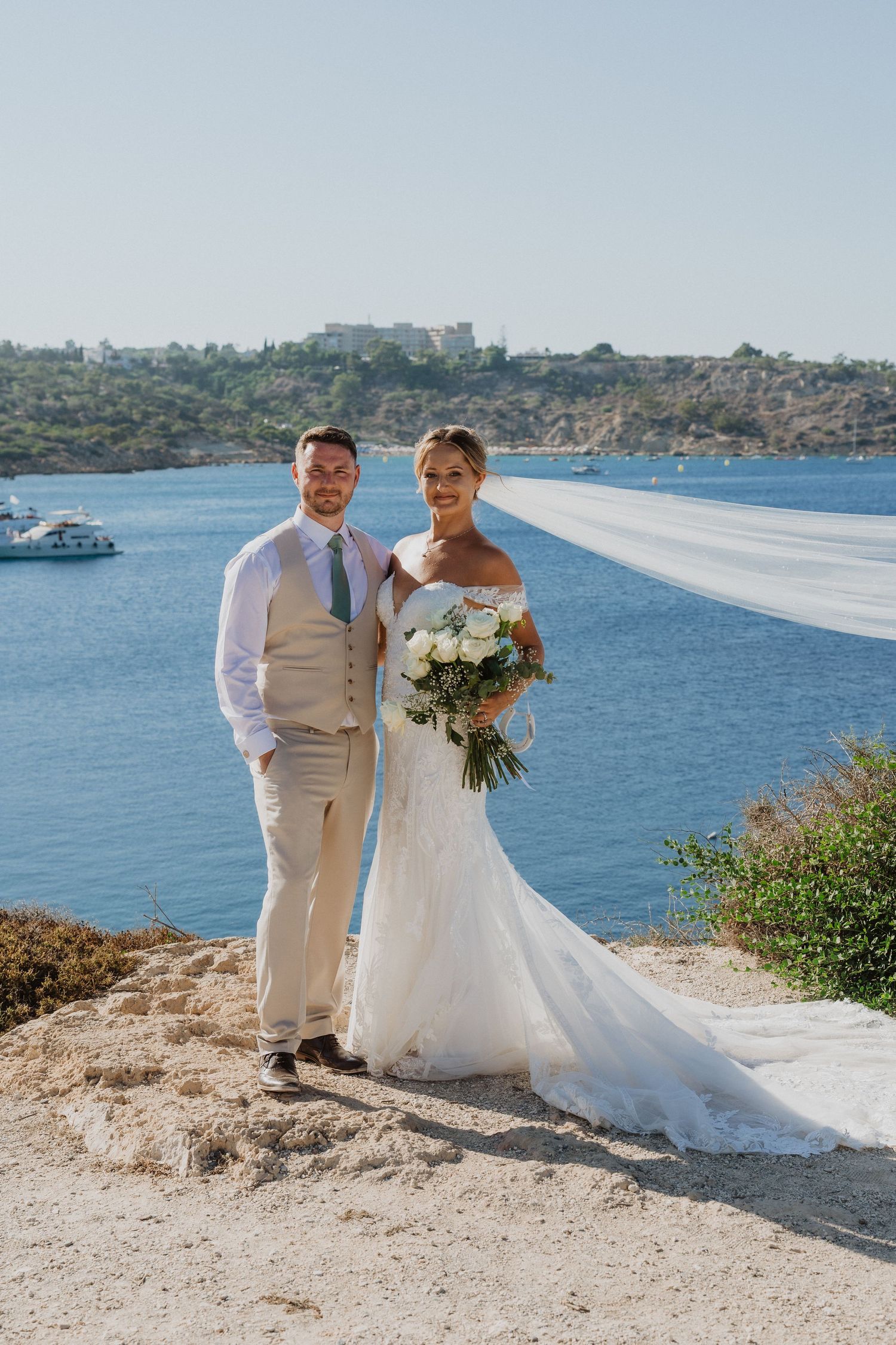 A couple poses for wedding photos along a scenic coastal cliffside with blue waters and rocky terrain in the background.