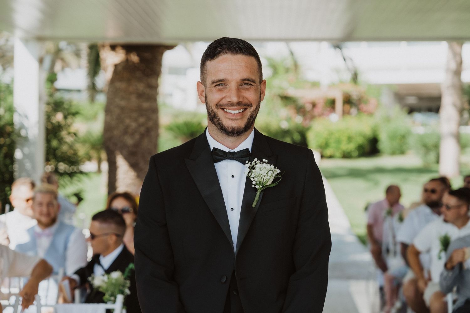 Groom in black tuxedo and bowtie smiles during outdoor wedding ceremony.