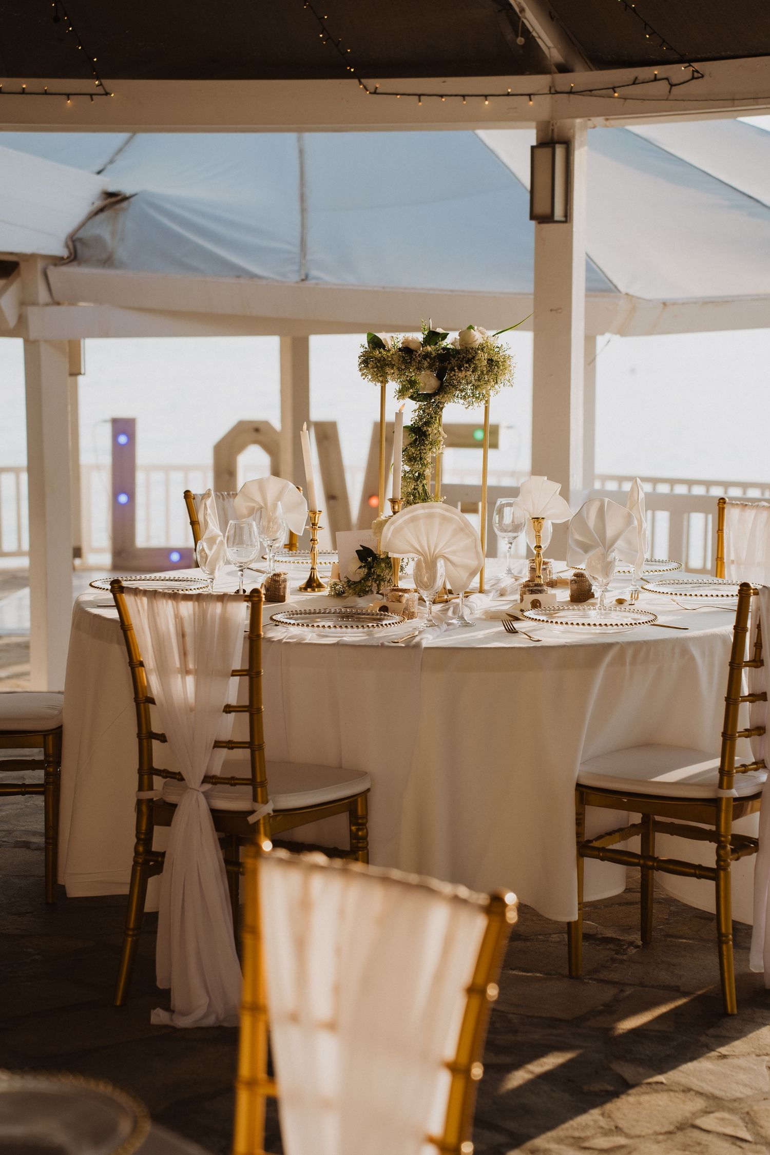 Romantic outdoor wedding reception table with gold chairs, white linens, floral centerpiece, and LOVE letters.