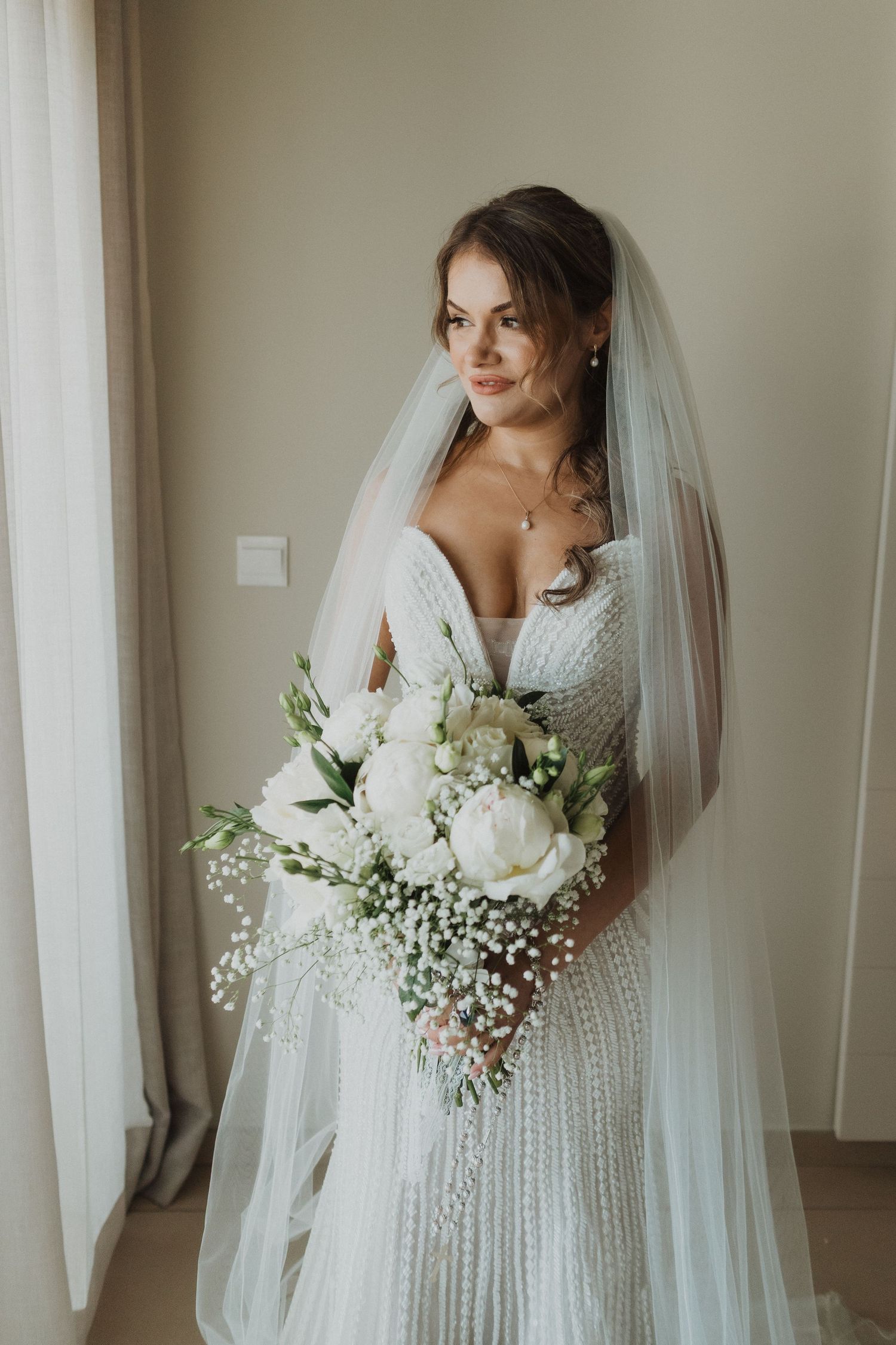 Elegant bride in beaded wedding gown holding white peony bouquet with veil indoors.