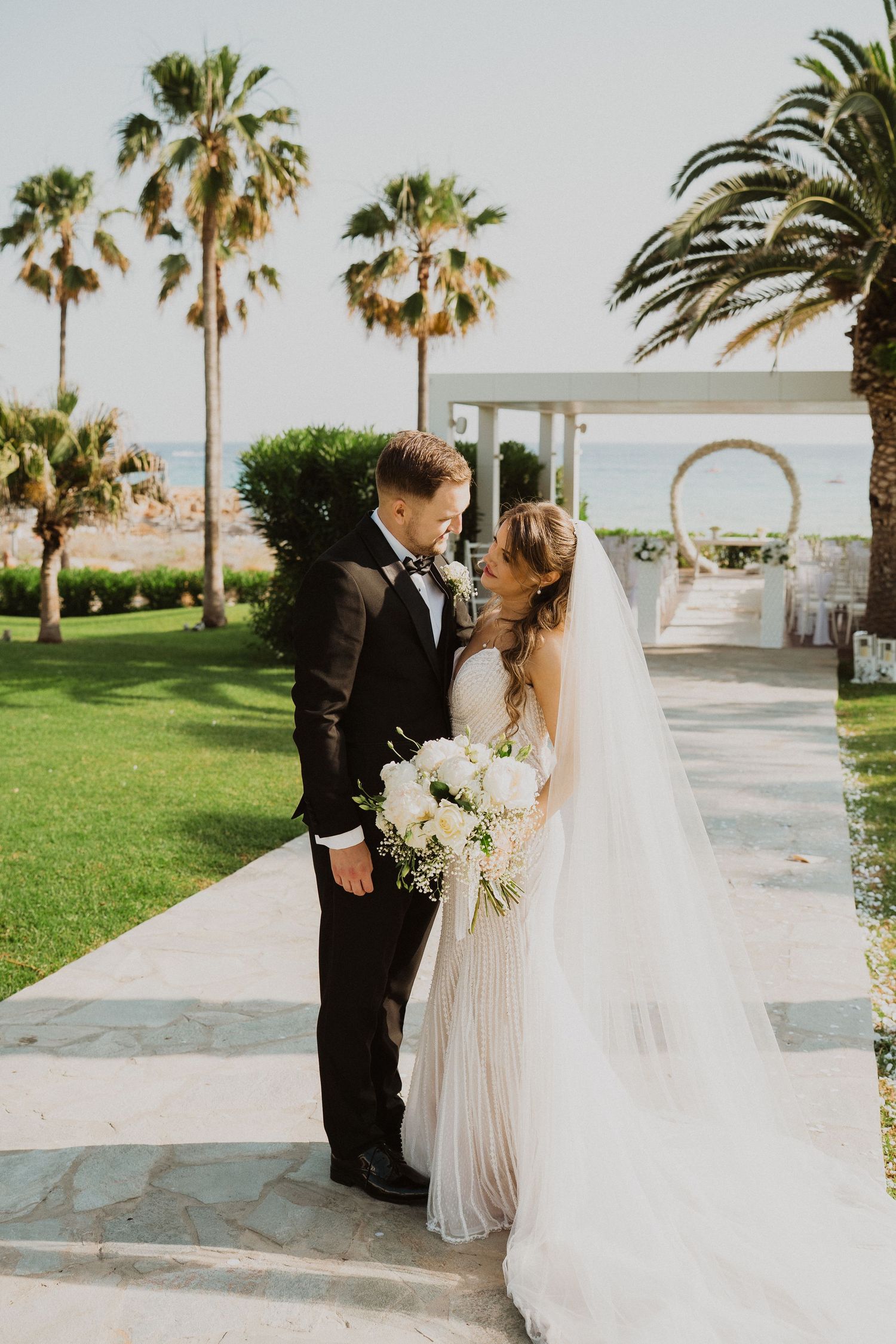 Bride and groom share intimate moment on garden path with palm trees and circular arch backdrop.