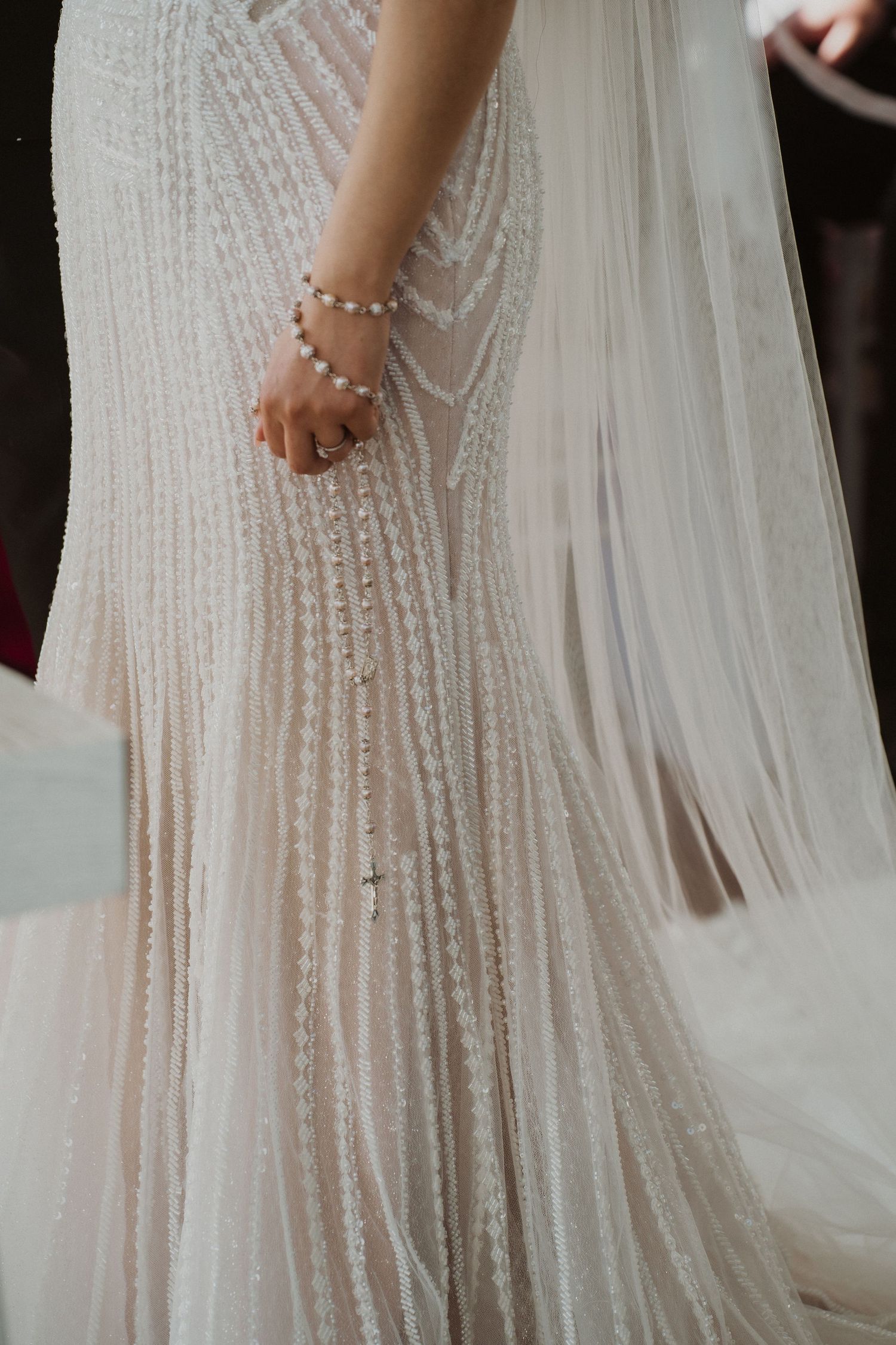 Close-up of a bride in a pearl-beaded wedding dress holding a rosary bracelet with a flowing veil.