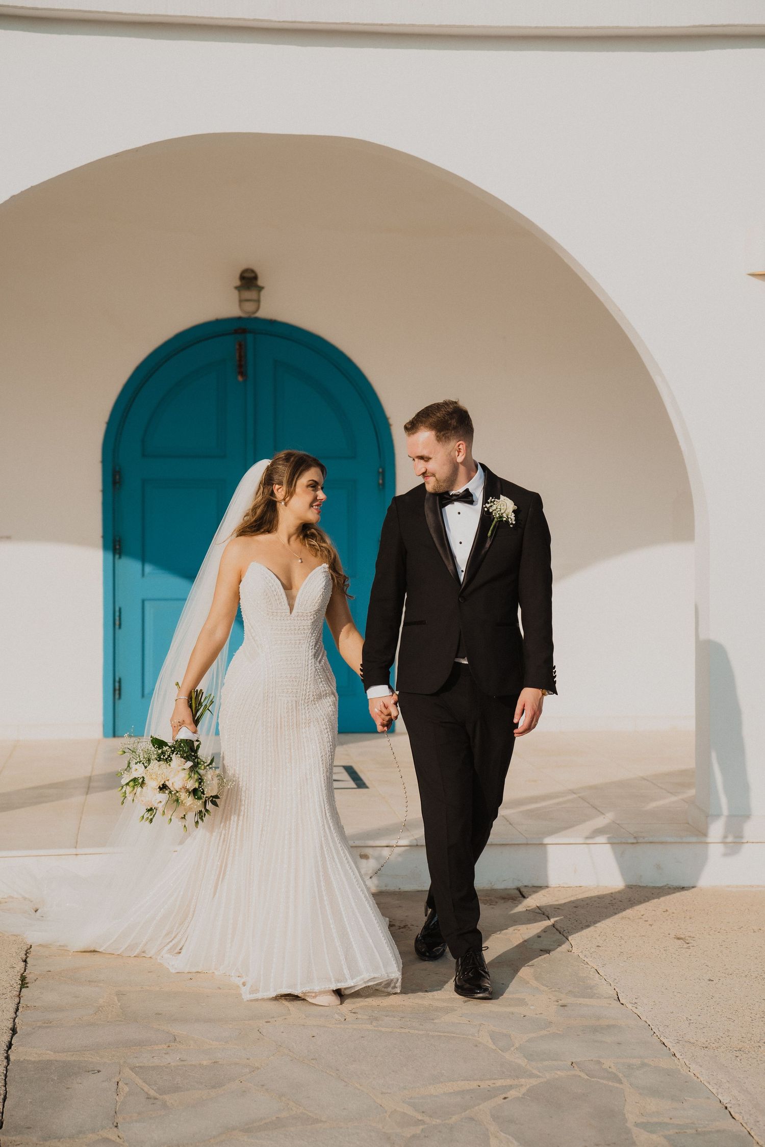 Bride in fitted lace gown and groom in black tuxedo walking hand in hand under white arches with teal door.