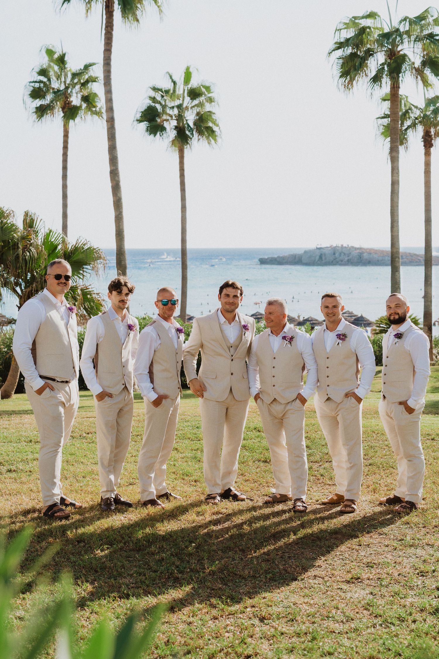 Groomsmen in cream colored suits line up on grass with palm trees and ocean in background.