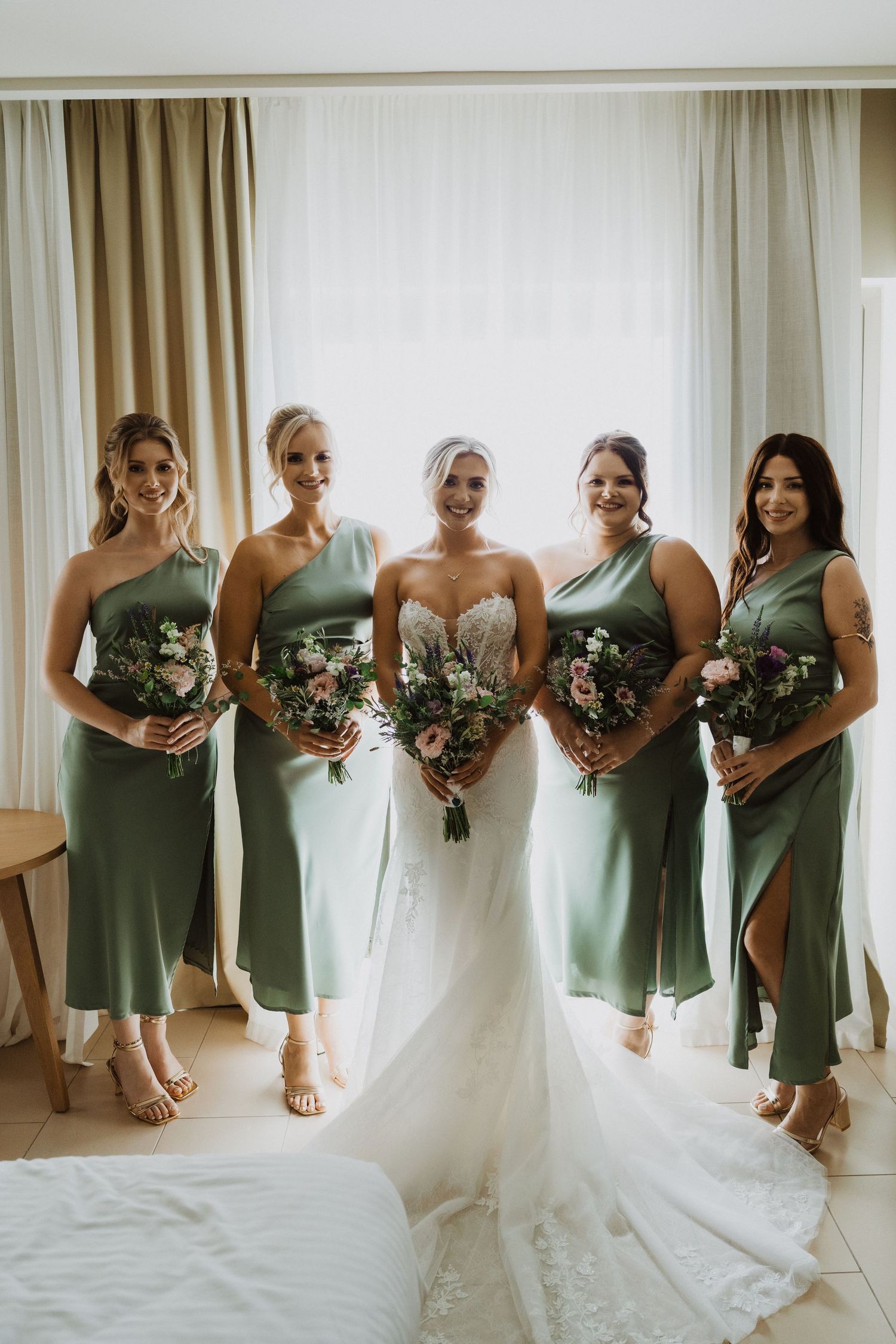 Wedding party in sage green bridesmaid dresses holding floral bouquets lined up by window.