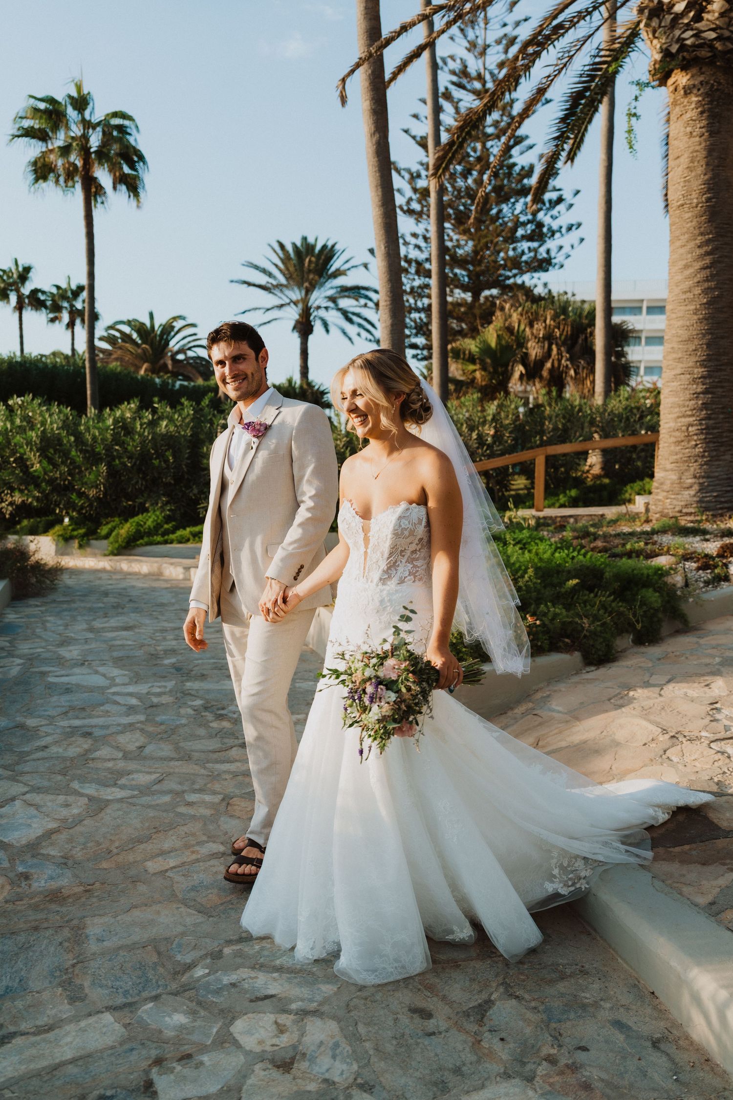 Bride in flowing white gown and groom walk through tropical garden path with palm trees.