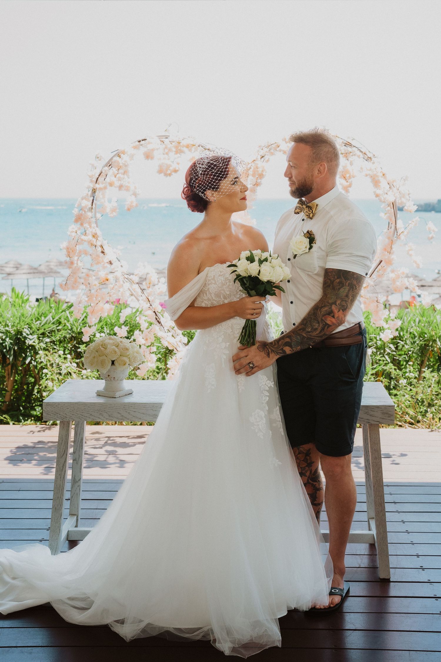 A beach wedding couple poses under a floral arch with the ocean in the background.