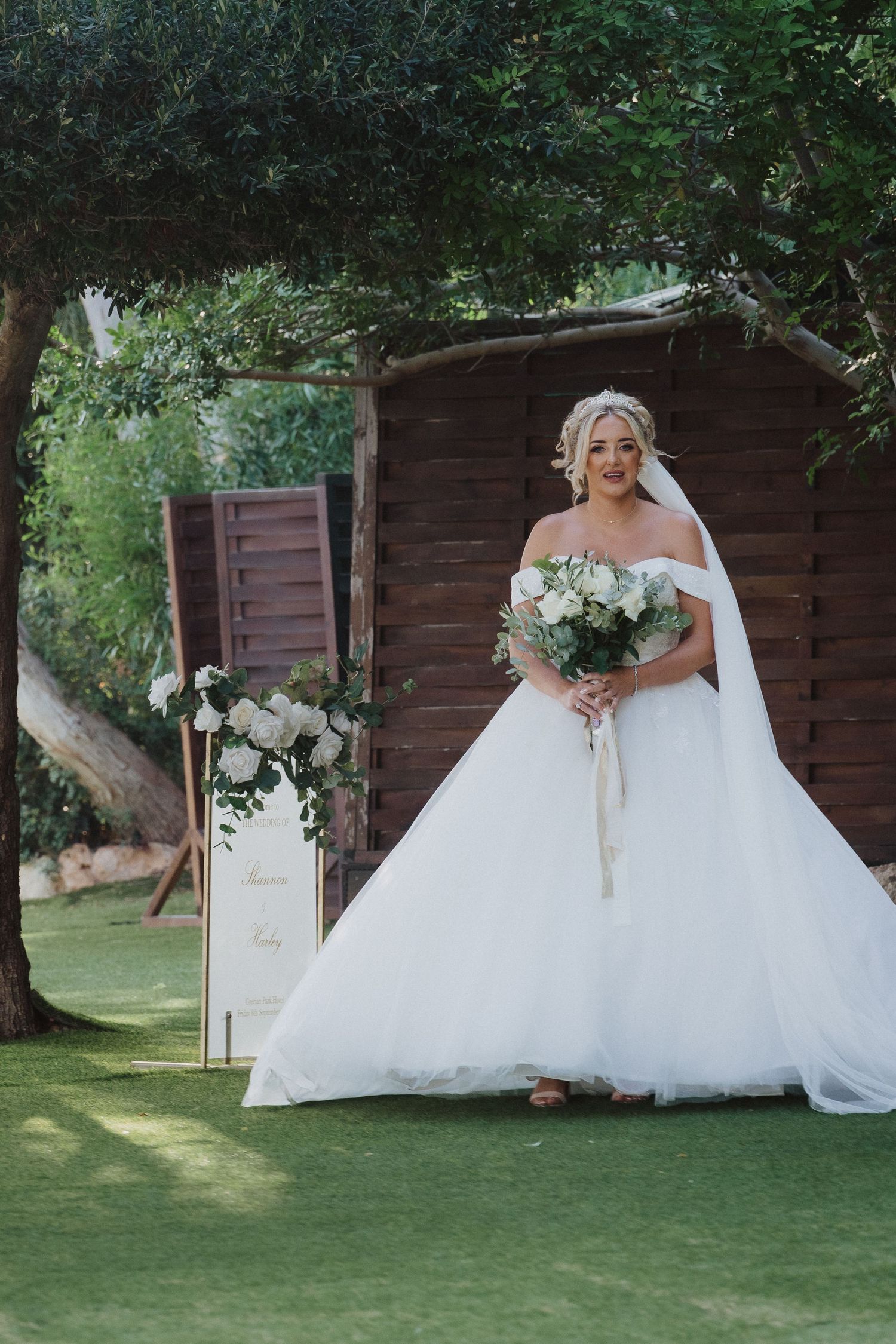 A wedding portrait showing a bride in a white ballgown holding a bouquet in front of a rustic wooden structure.