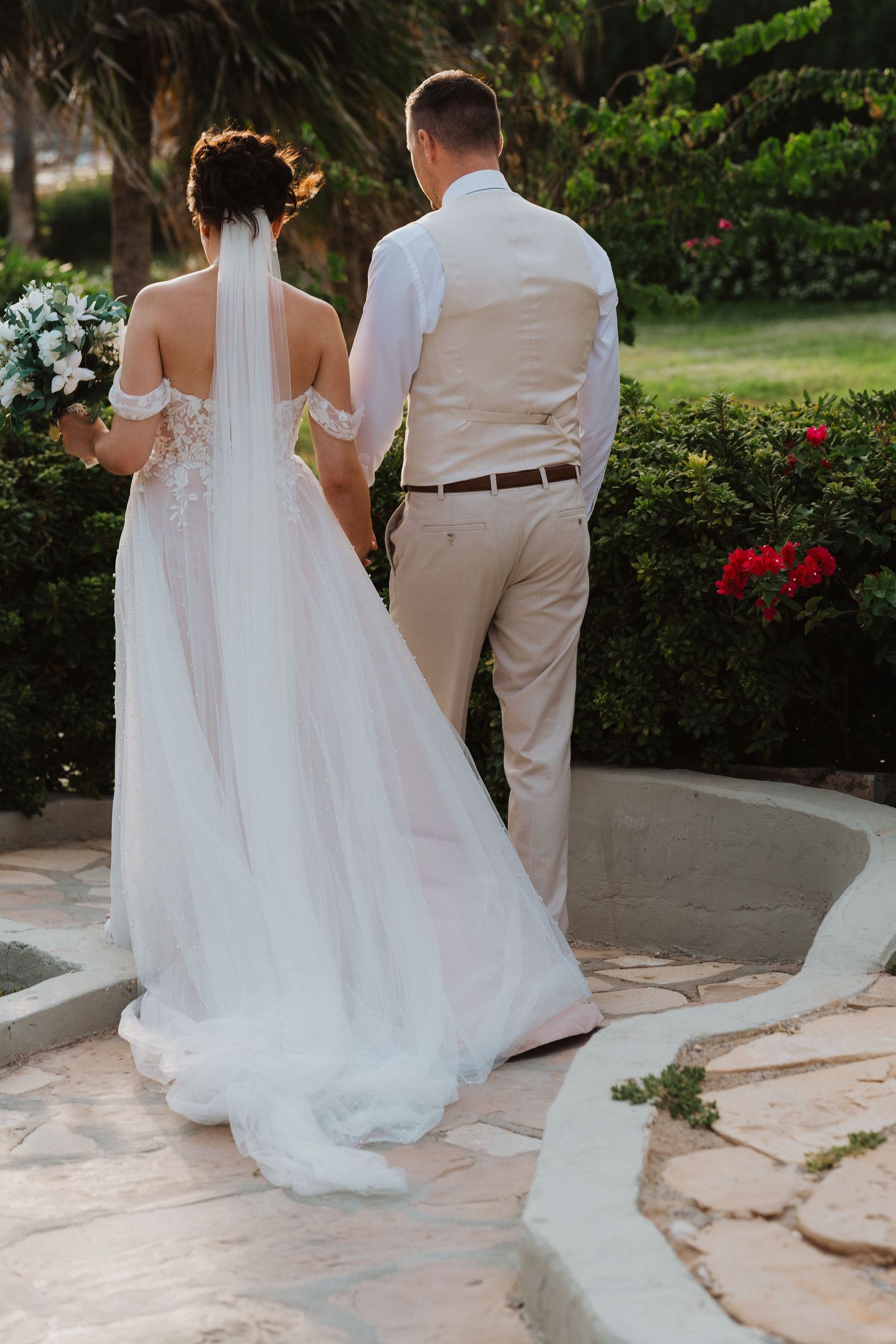 A couple in wedding attire stand on a curved garden path during golden hour, with the bride in a flowing white dress and veil.