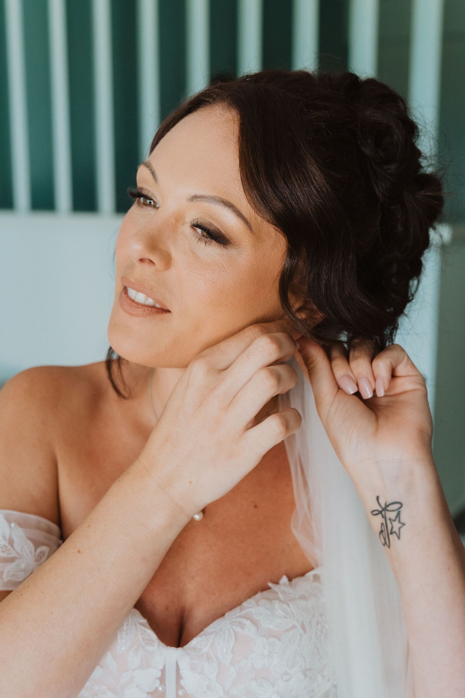 Bride in white lace dress puts on earrings while looking up with gentle smile, small butterfly tattoo visible on wrist.