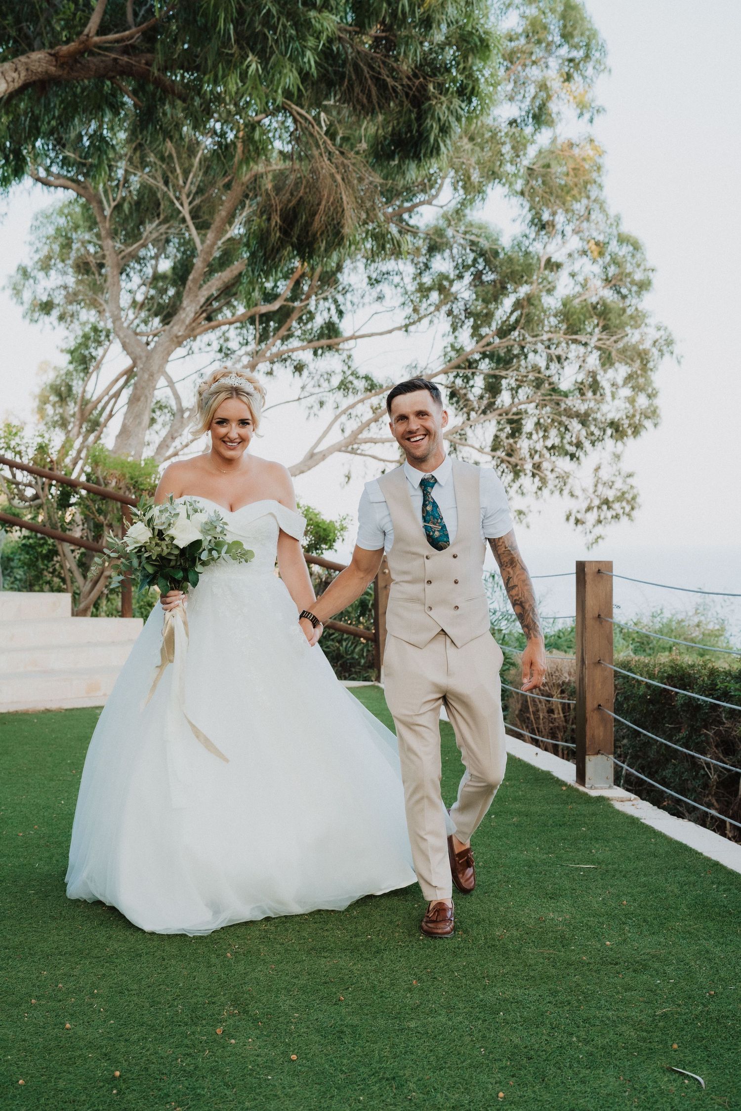 A couple walks together on grass with eucalyptus trees and ocean views in the background.