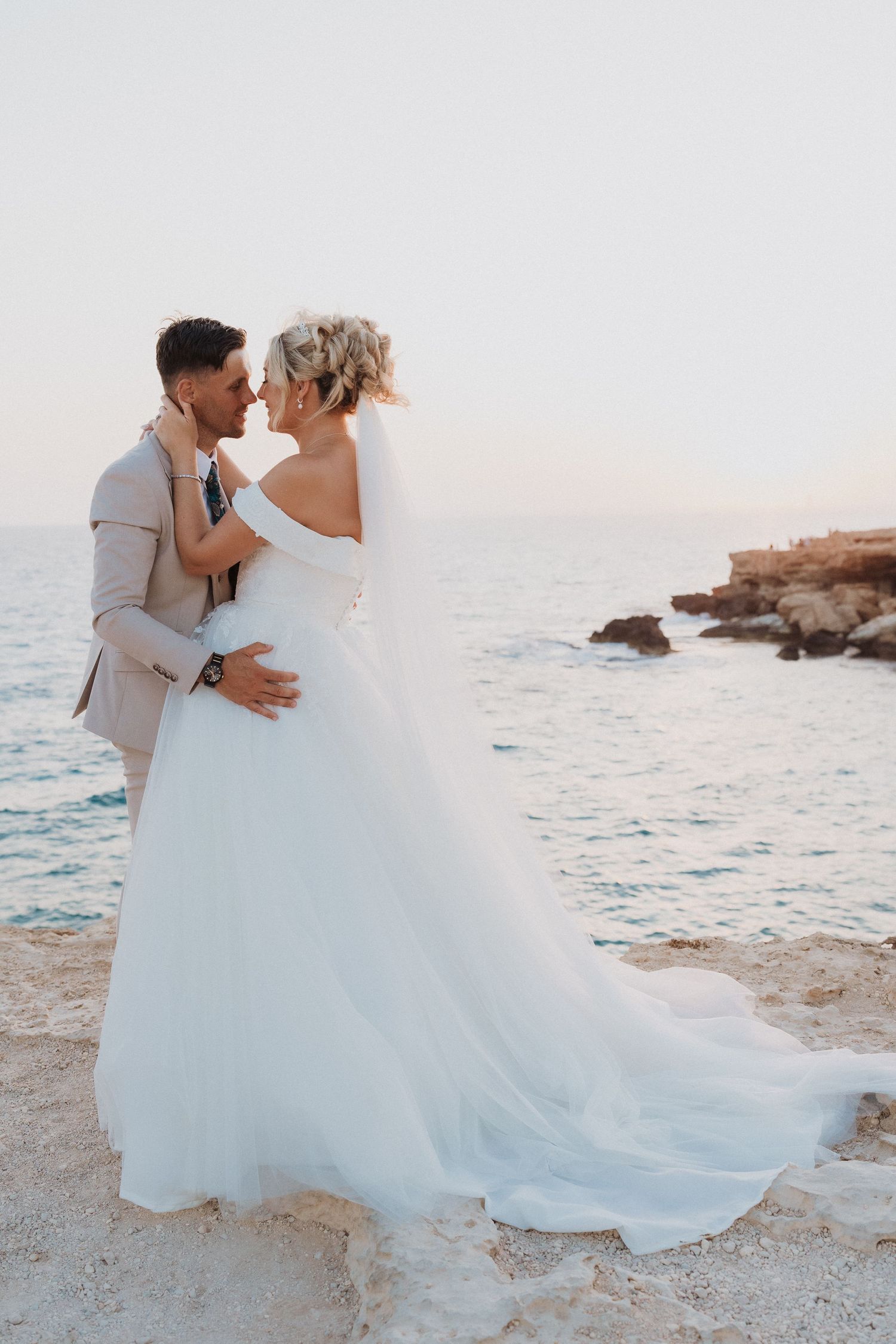 Romantic wedding couple embraces on rocky coastal cliff at sunset with bride wearing flowing white gown.