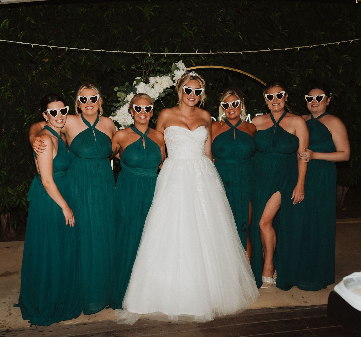 A group wearing coordinated emerald green dresses and sunglasses pose together at a wedding celebration.