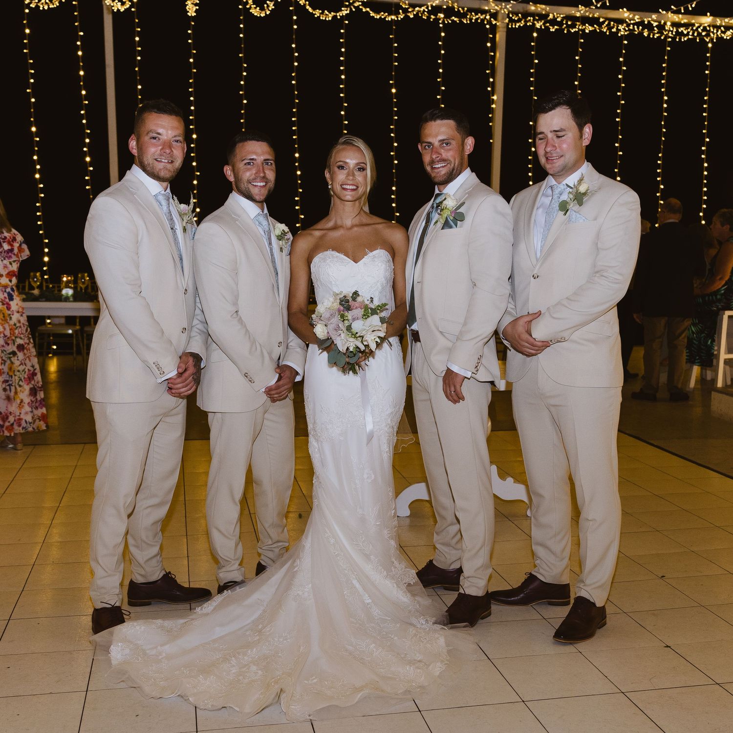 Wedding party in beige suits and white gown poses under string lights at reception.