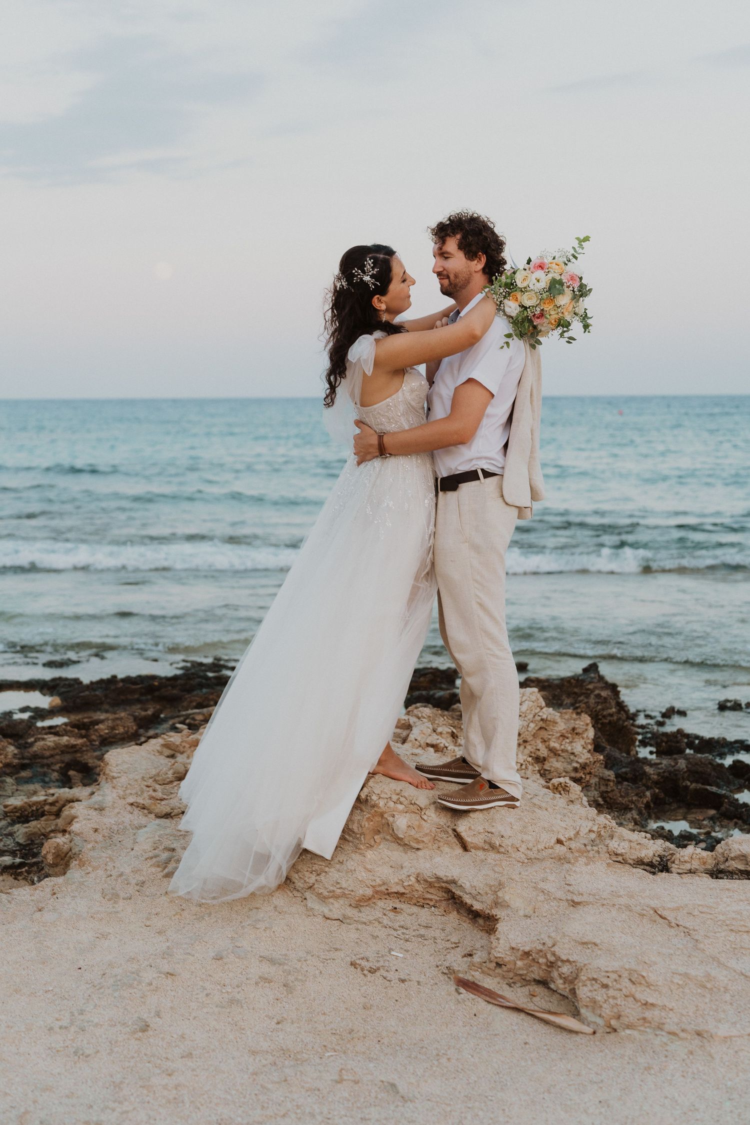 A romantic embrace on rocky coastal terrain during a beach wedding ceremony.