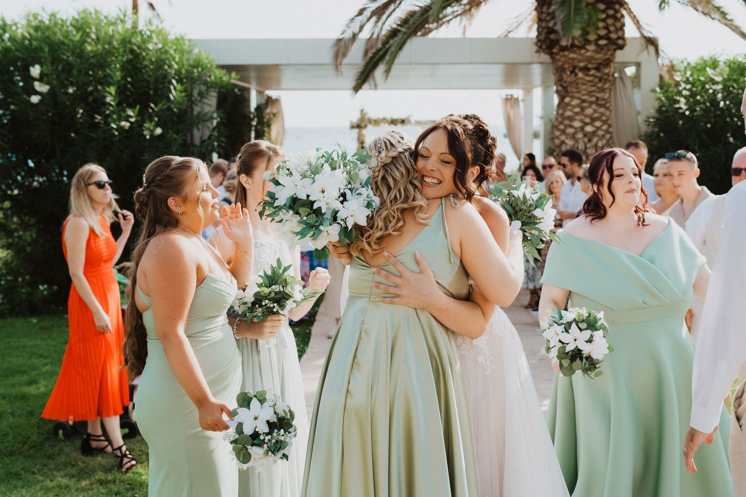 Bridesmaids in pastel mint and sage dresses laugh together while holding white floral bouquets at an outdoor wedding.