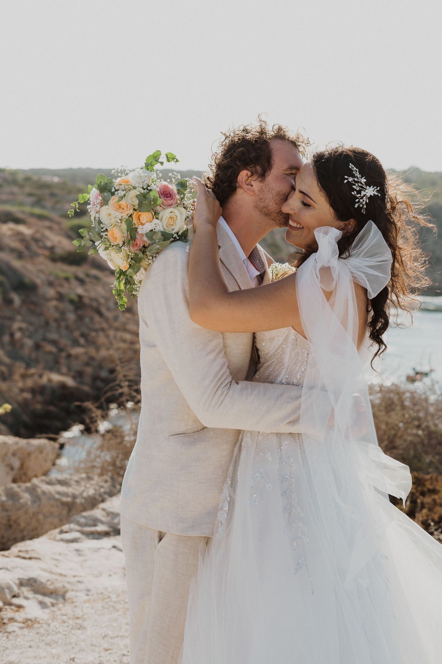 A tender wedding moment captured against a backdrop of Mediterranean waters and rocky coastline.