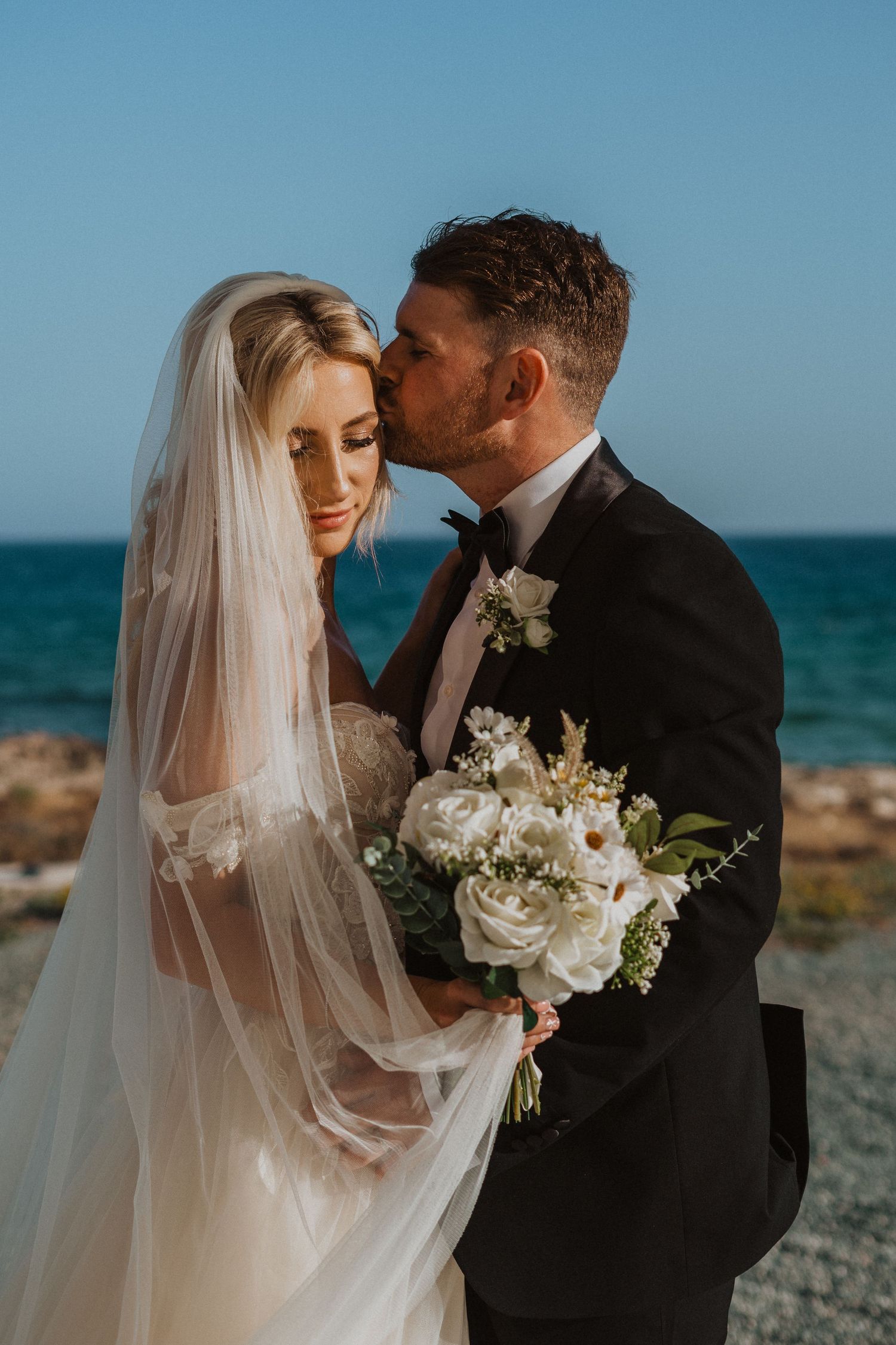 Tender moment between newlyweds on coastal backdrop with white rose bouquet.