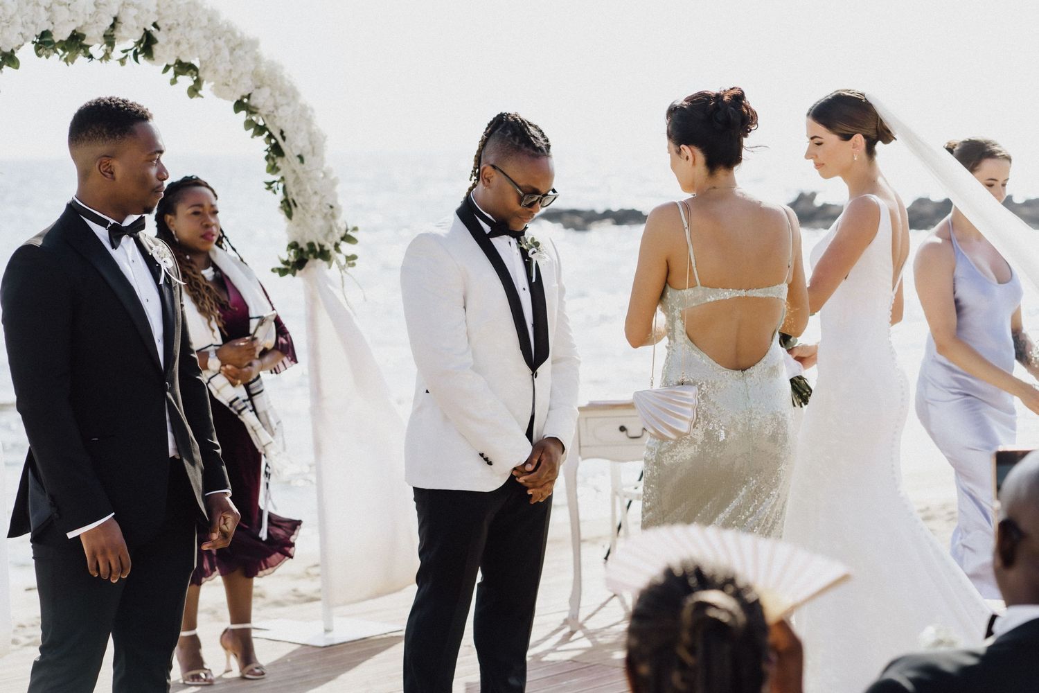 Wedding party gathered under a floral arch during a beachside ceremony.