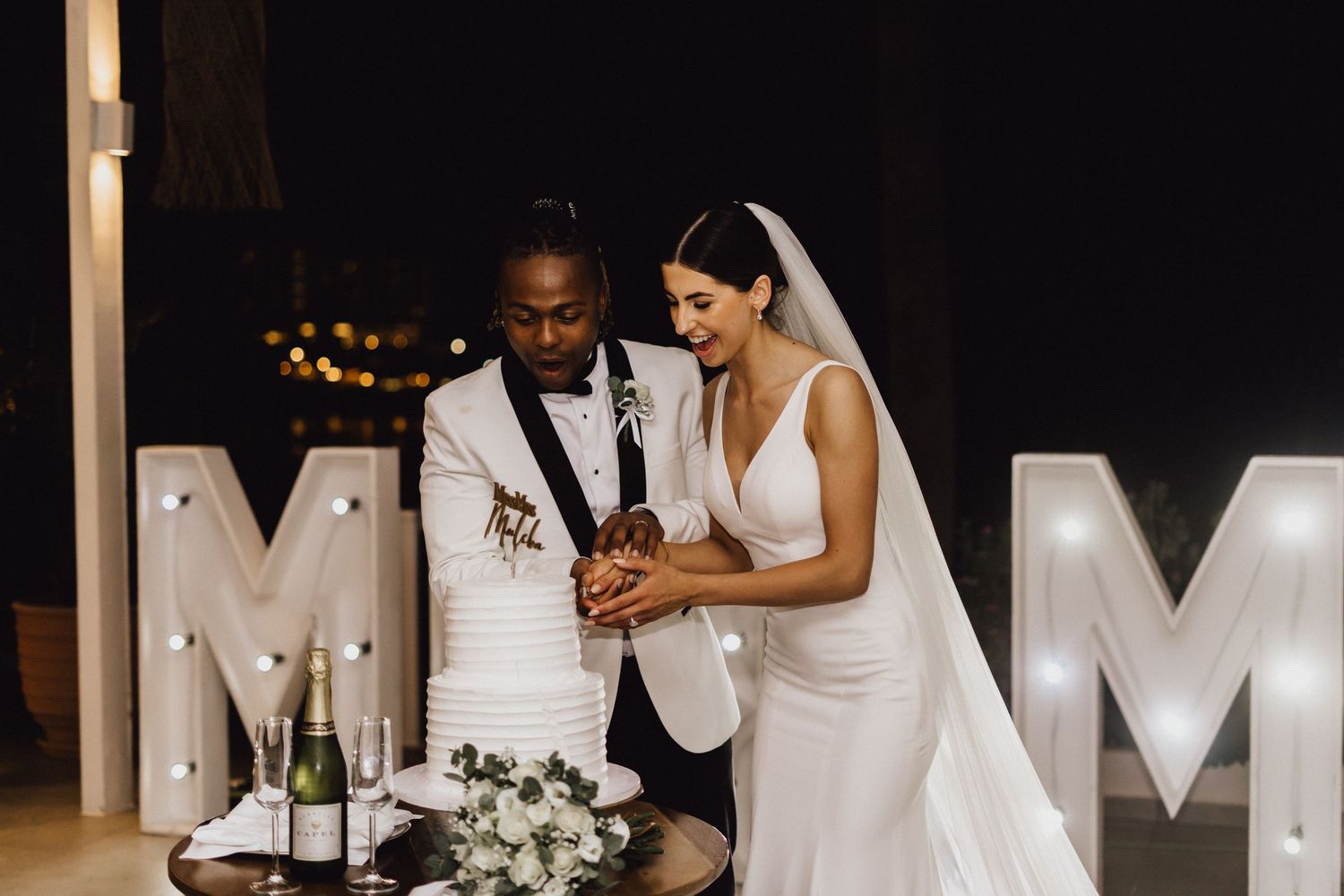 Newlyweds cutting their wedding cake beside illuminated marquee letters.