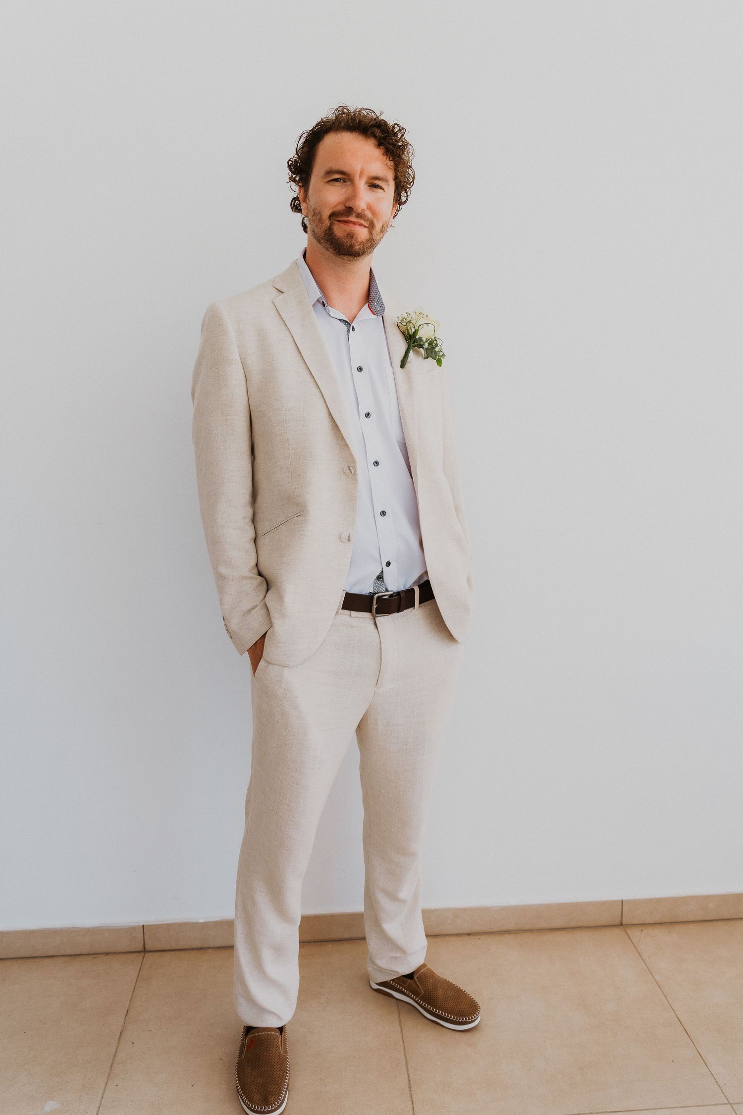 Groom in beige suit with boutonniere poses against white wall at wedding.