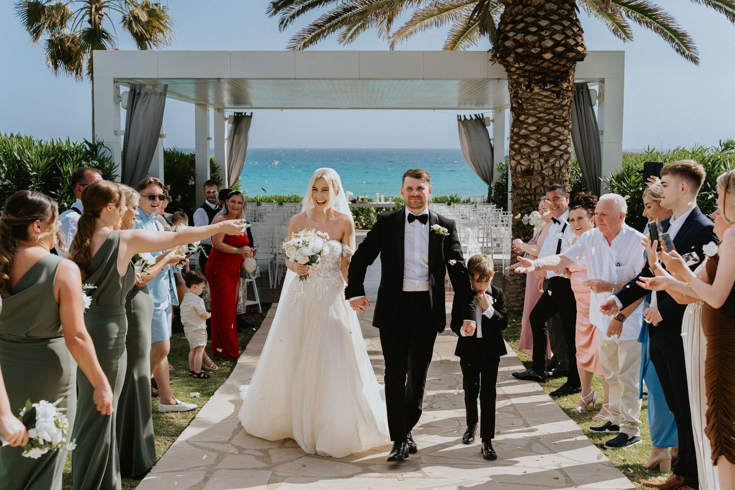 A wedding recessional along a palm-lined walkway with guests cheering and the ocean in view.