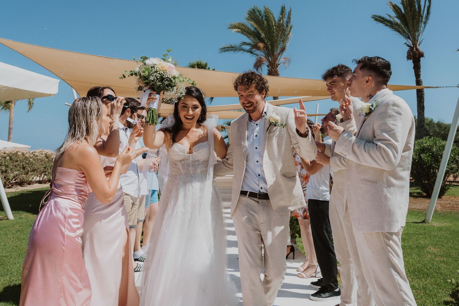 Wedding party celebrates on palm-lined pathway under tan shade sails at beach resort.
