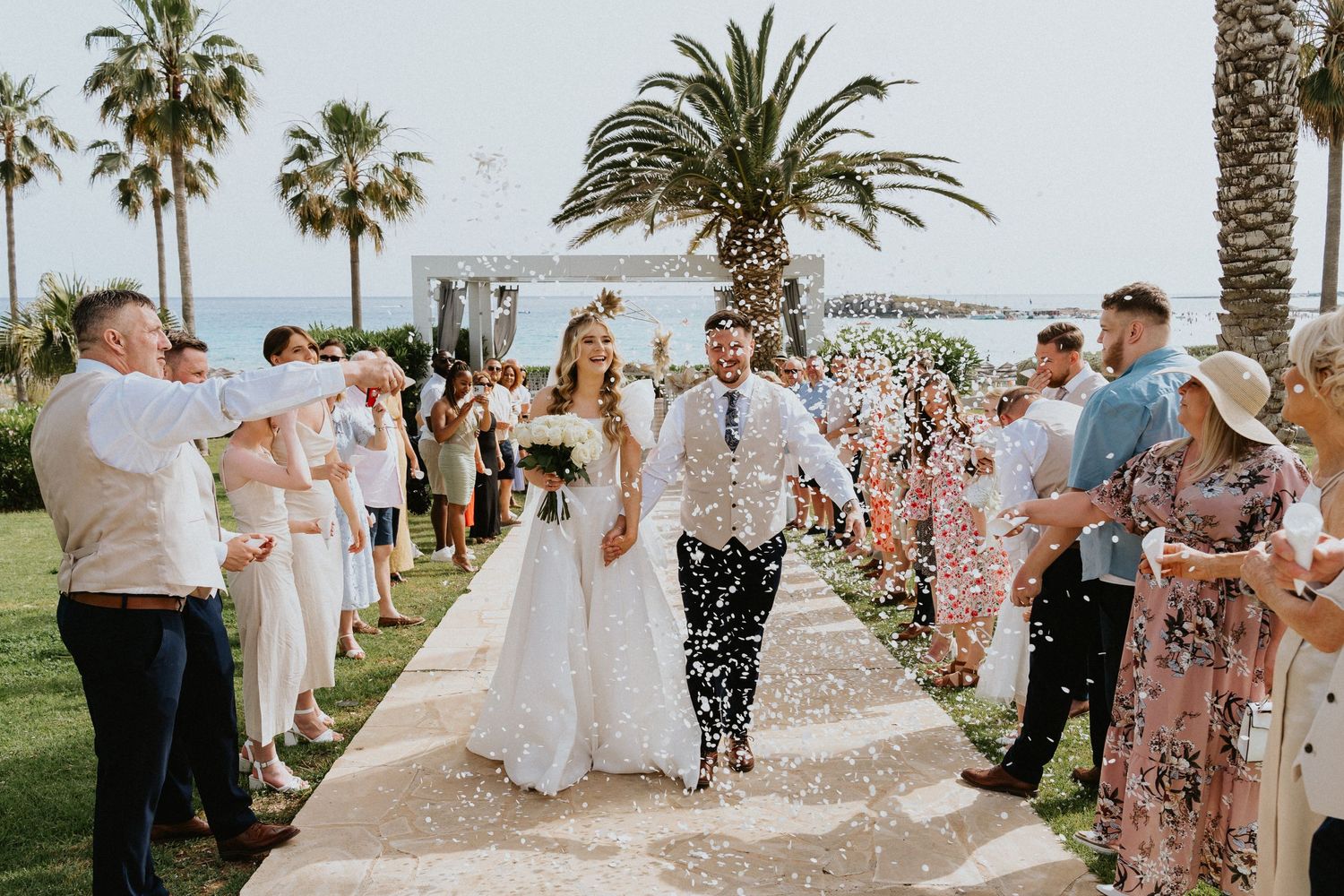 Guests shower confetti on newlyweds walking down palm-lined oceanfront path.