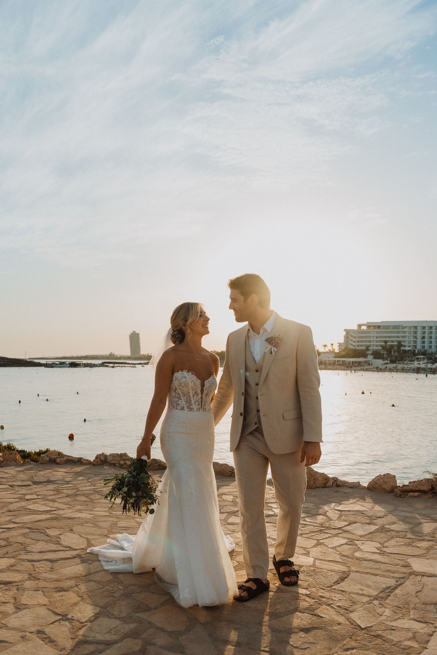 Newlyweds stand together on a stone terrace during sunset with ocean views.