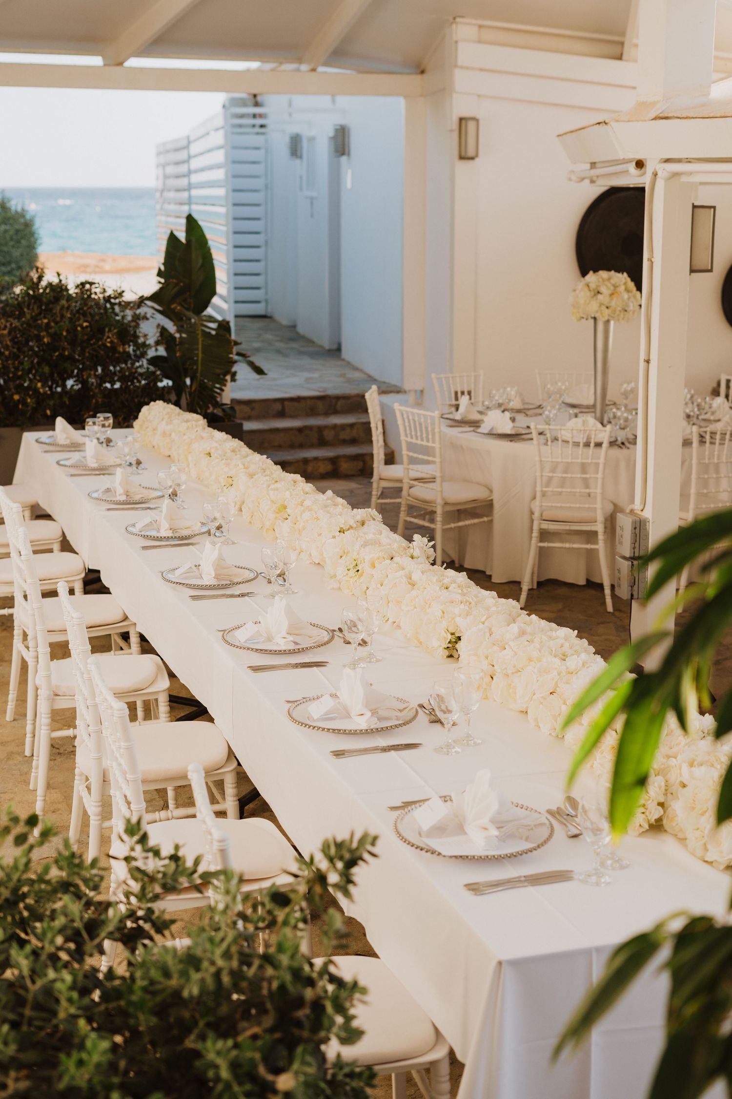 Elegant white wedding reception tables set with place settings and floral arrangements in a bright indoor venue.