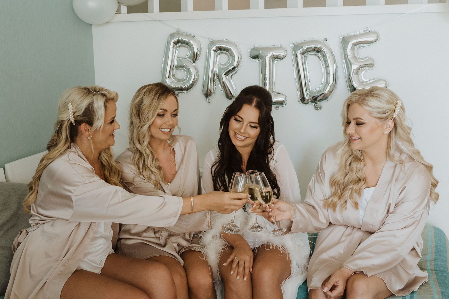 Wedding party in matching robes toasting with champagne beneath silver BRIDE balloons.