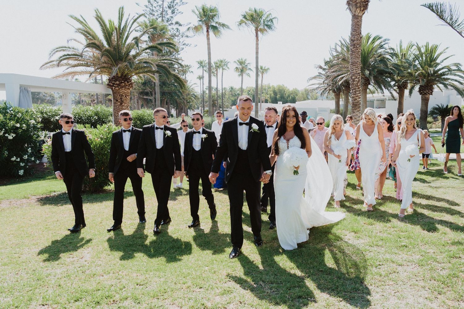 Wedding party and guests gathered on a grassy area with tropical palm trees.