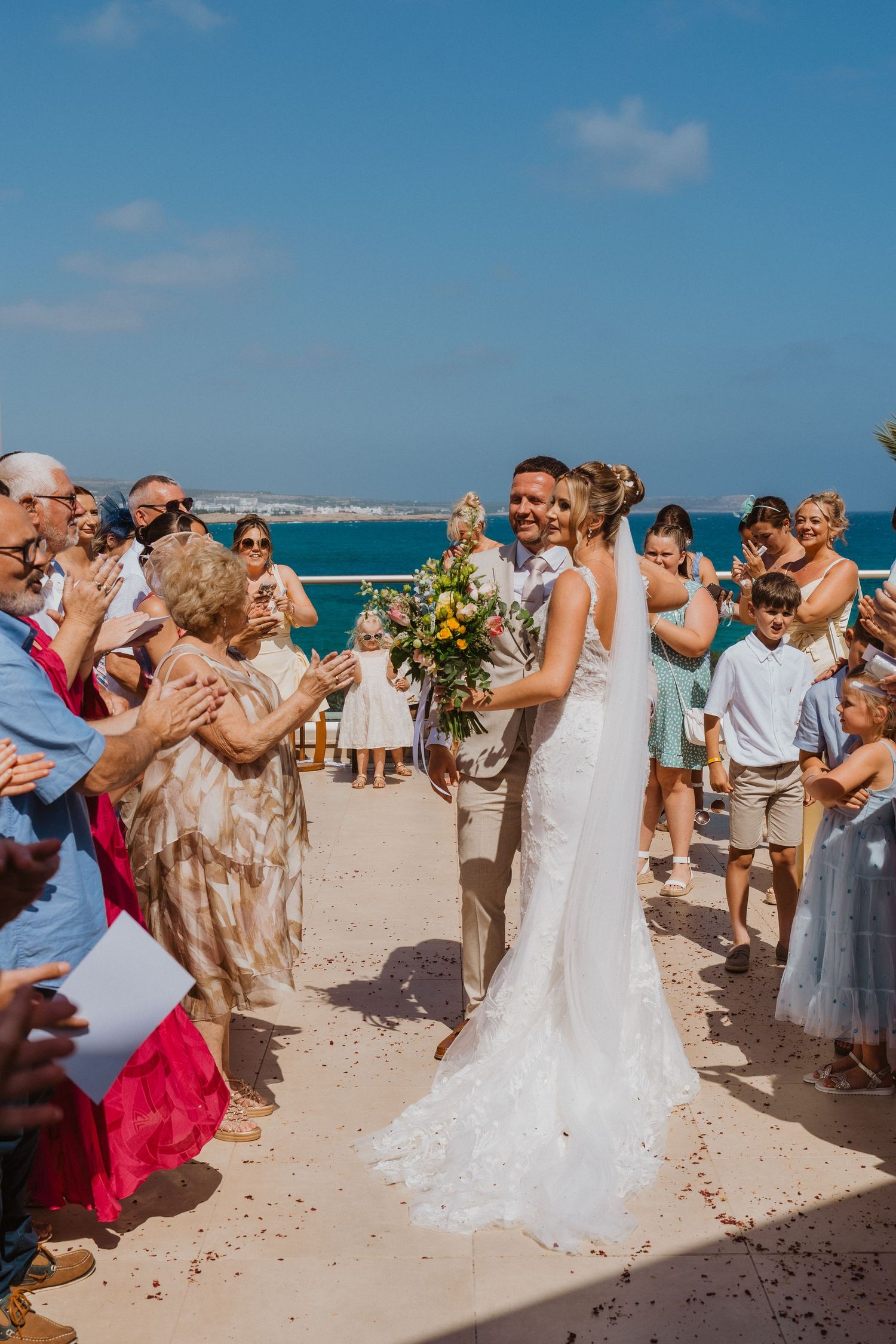 Wedding guests form a celebratory tunnel on a sunny beach while throwing flower petals as newlyweds walk through.