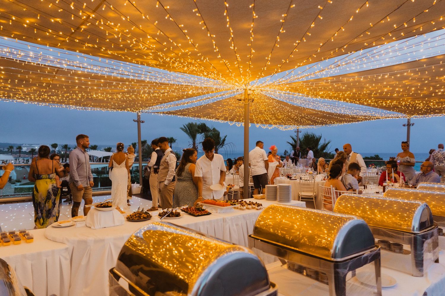 Outdoor evening buffet reception under glowing string lights with ocean view and guests gathering around serving stations.