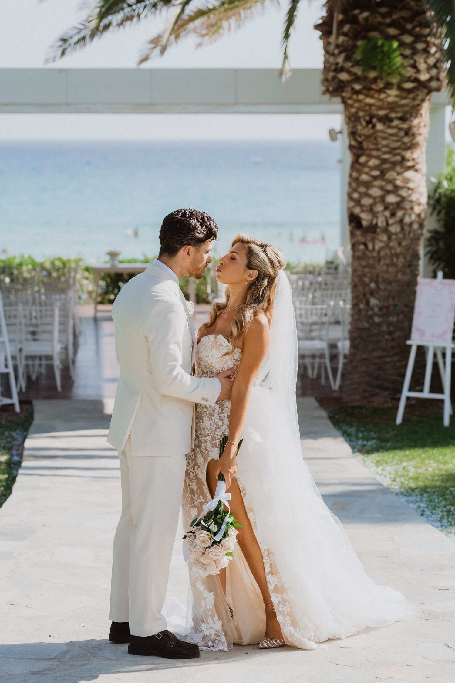 A romantic wedding couple shares intimate moments in a tropical beachside venue with palm trees and white decor.