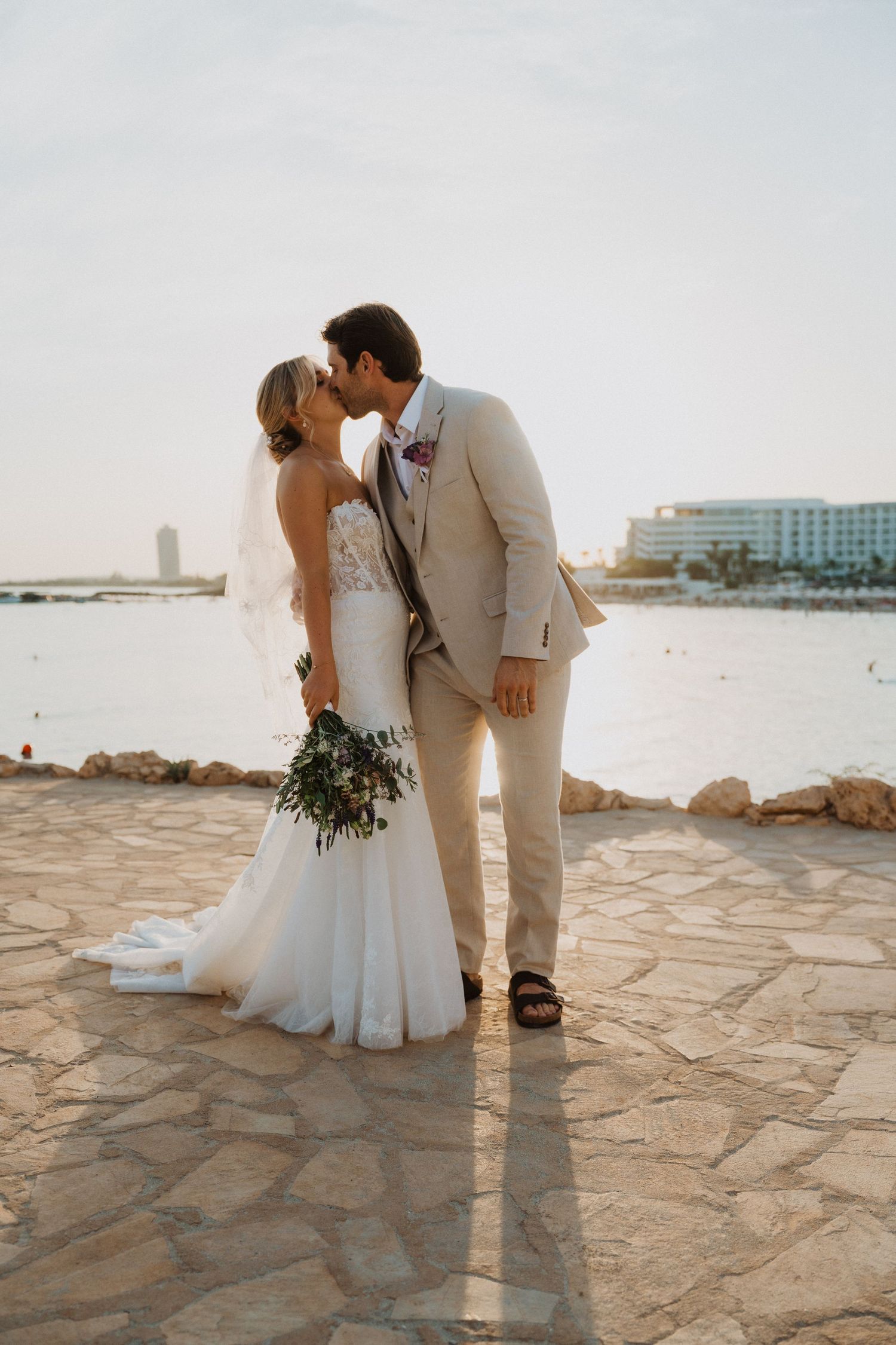 Wedding couple poses on seaside stone terrace during golden hour with waterfront buildings in background.