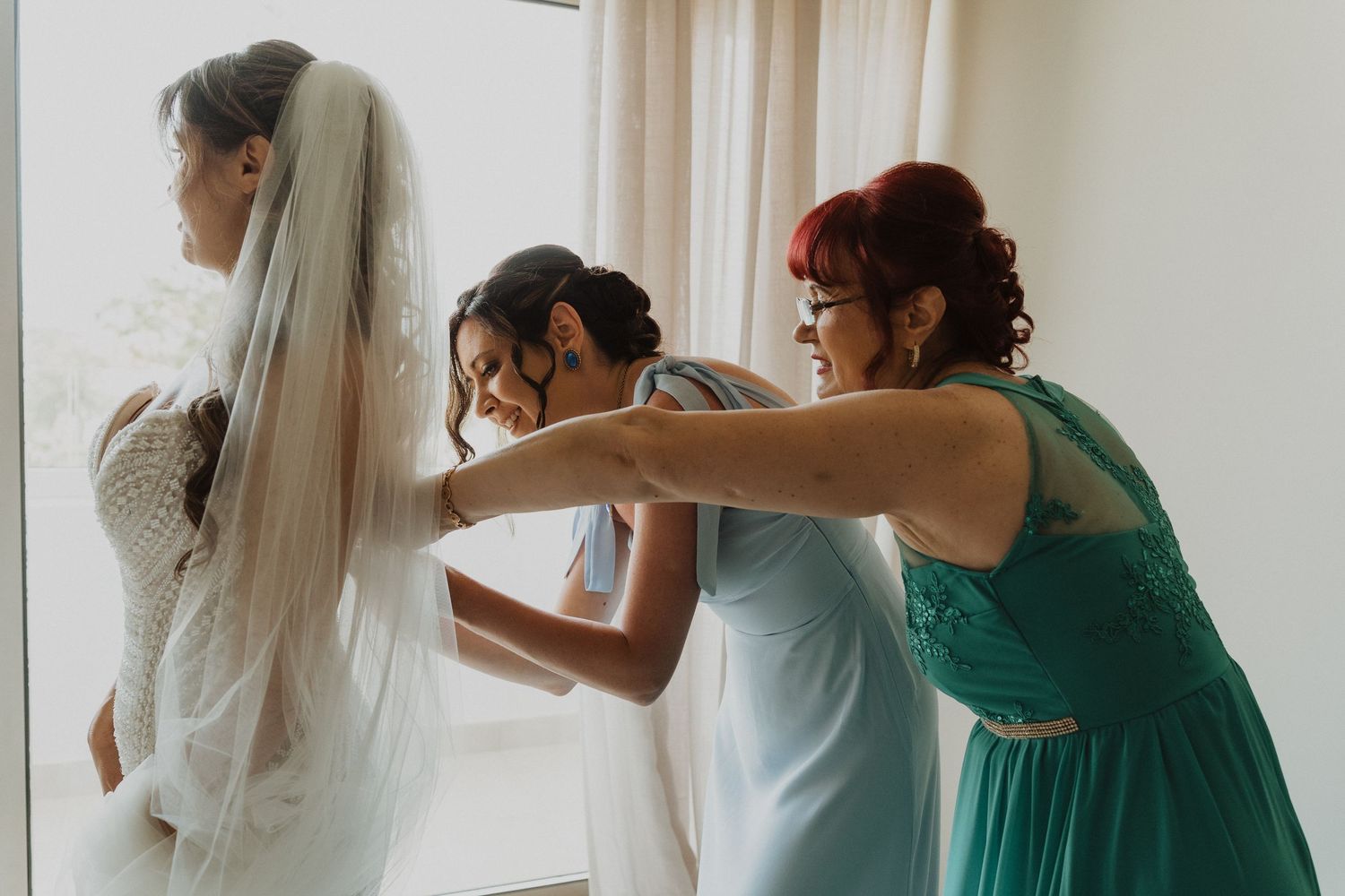 Bridesmaids helping bride adjust her wedding veil near a bright window before ceremony.