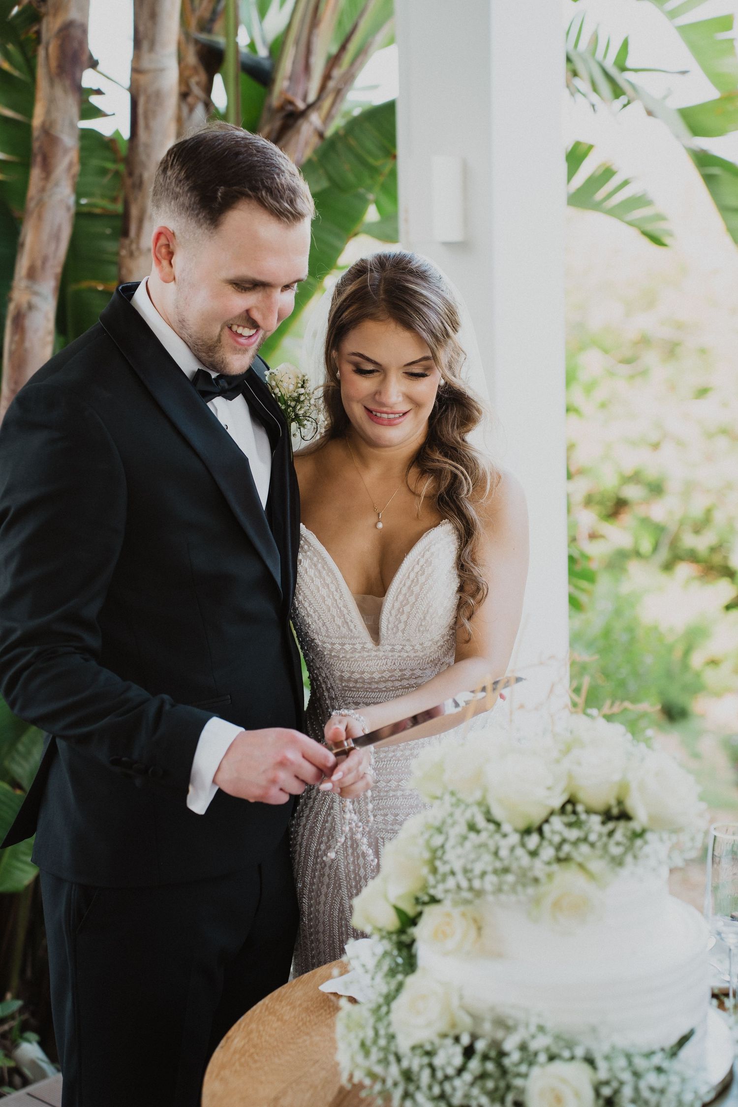 Smiling bride and groom cut their white floral wedding cake together at an outdoor reception.