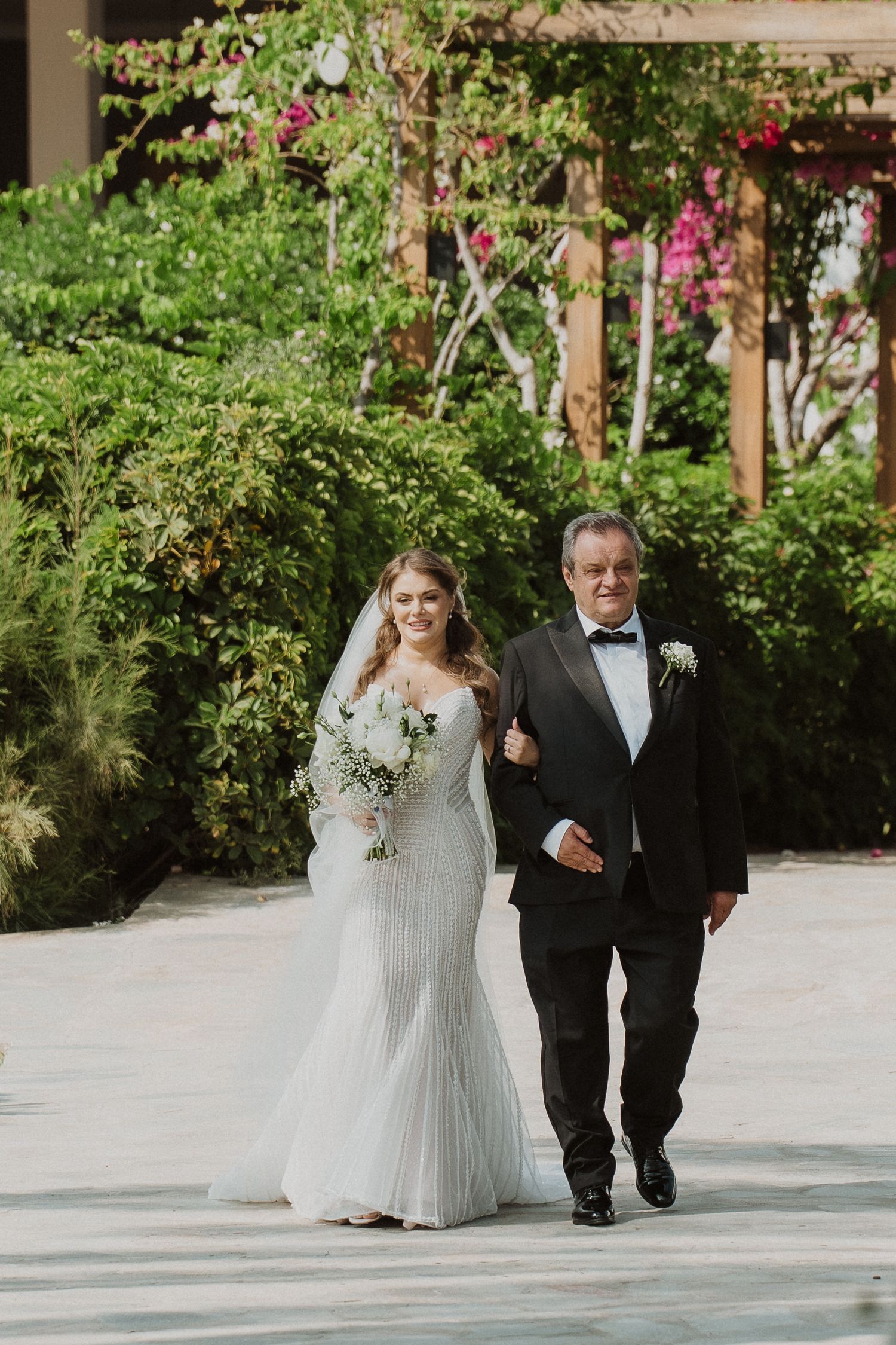 Bride in long-sleeve lace mermaid gown walks down outdoor aisle with escort holding white bouquet.