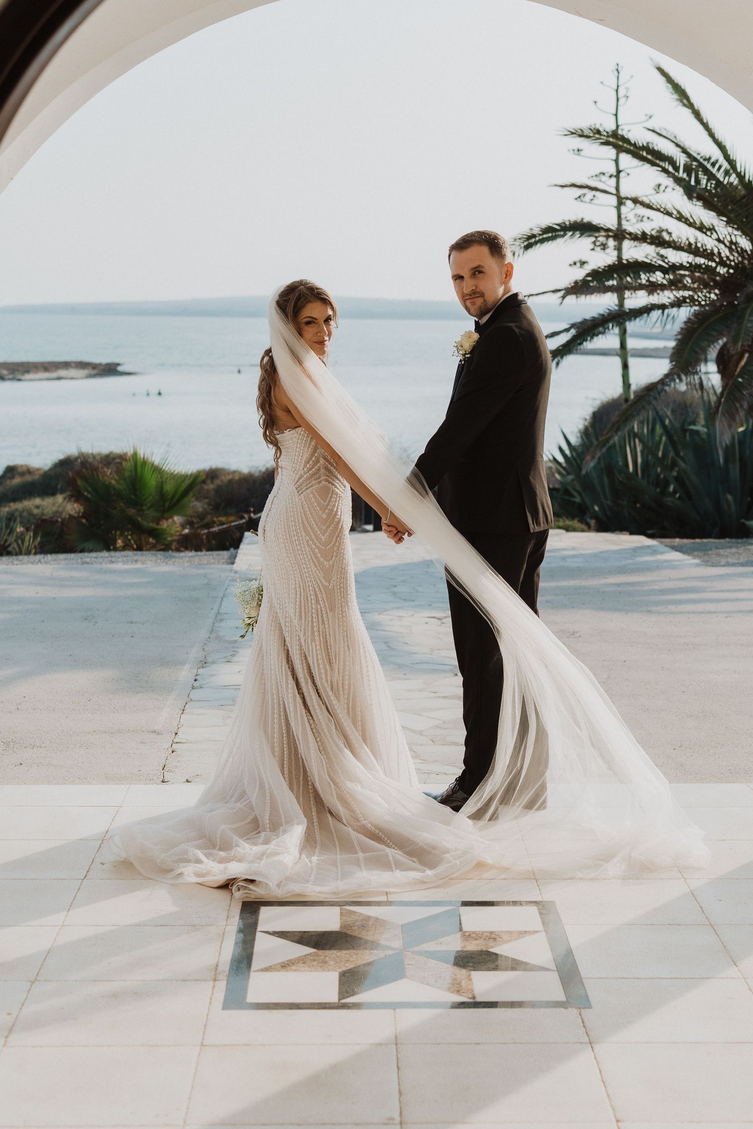 Bride in flowing veil and groom in tuxedo pose elegantly on terrace with sea views behind them.