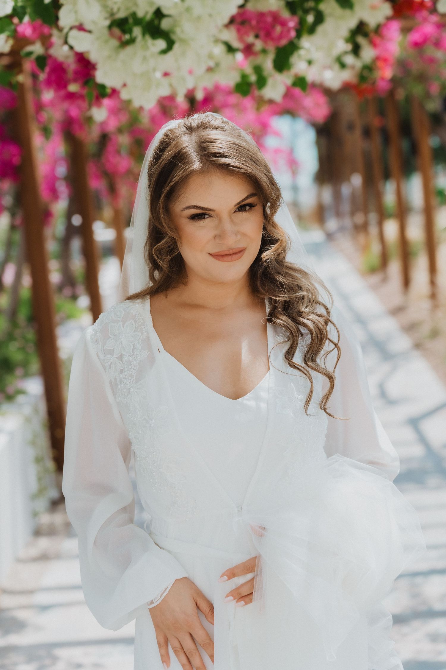 Bride in white lace long-sleeve gown smiling under pink bougainvillea arch on sunny outdoor pathway.