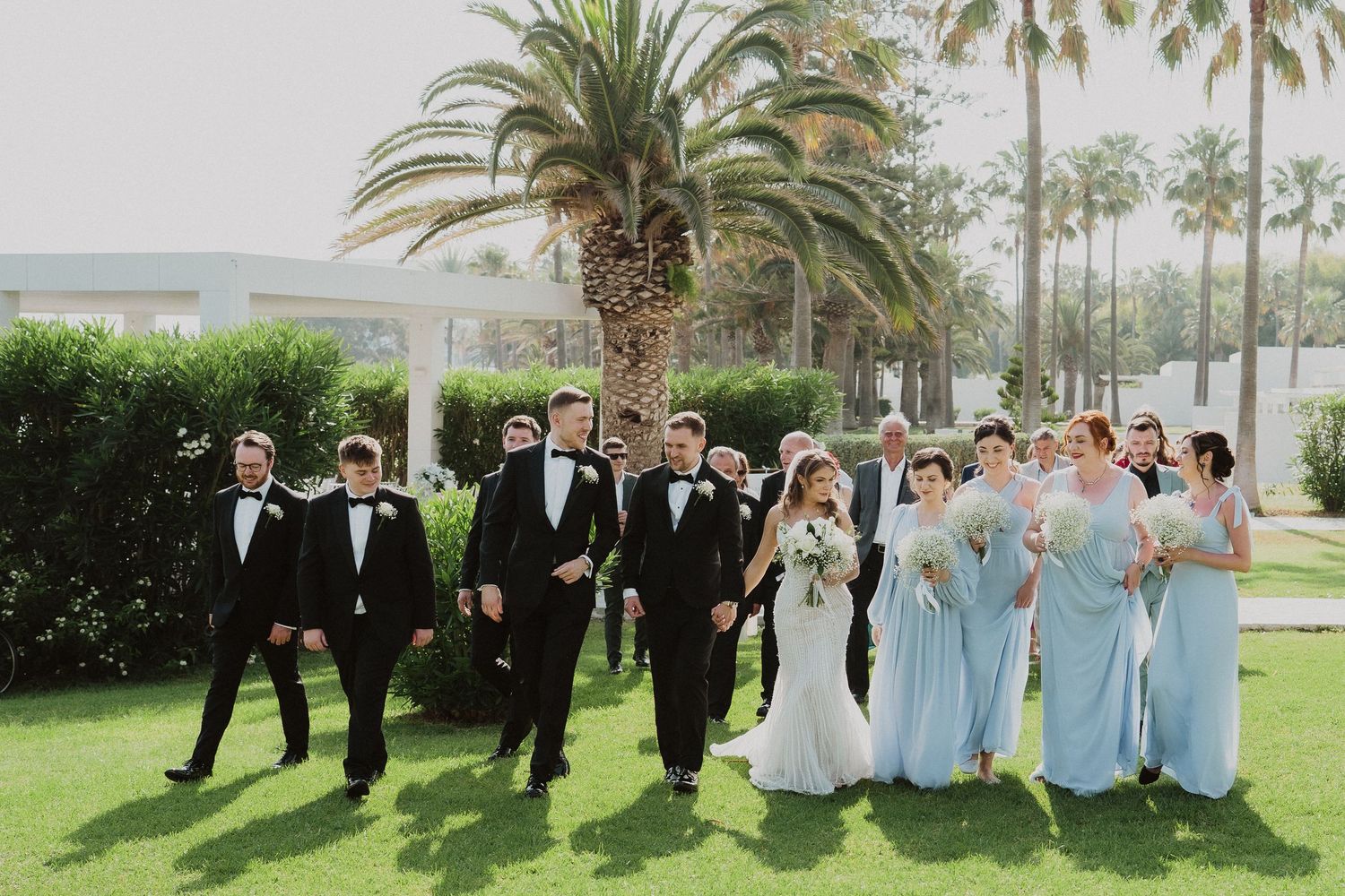 Wedding party walks outdoors near palm trees, groomsmen in tuxedos and bridesmaids in light blue gowns with bride and groom.