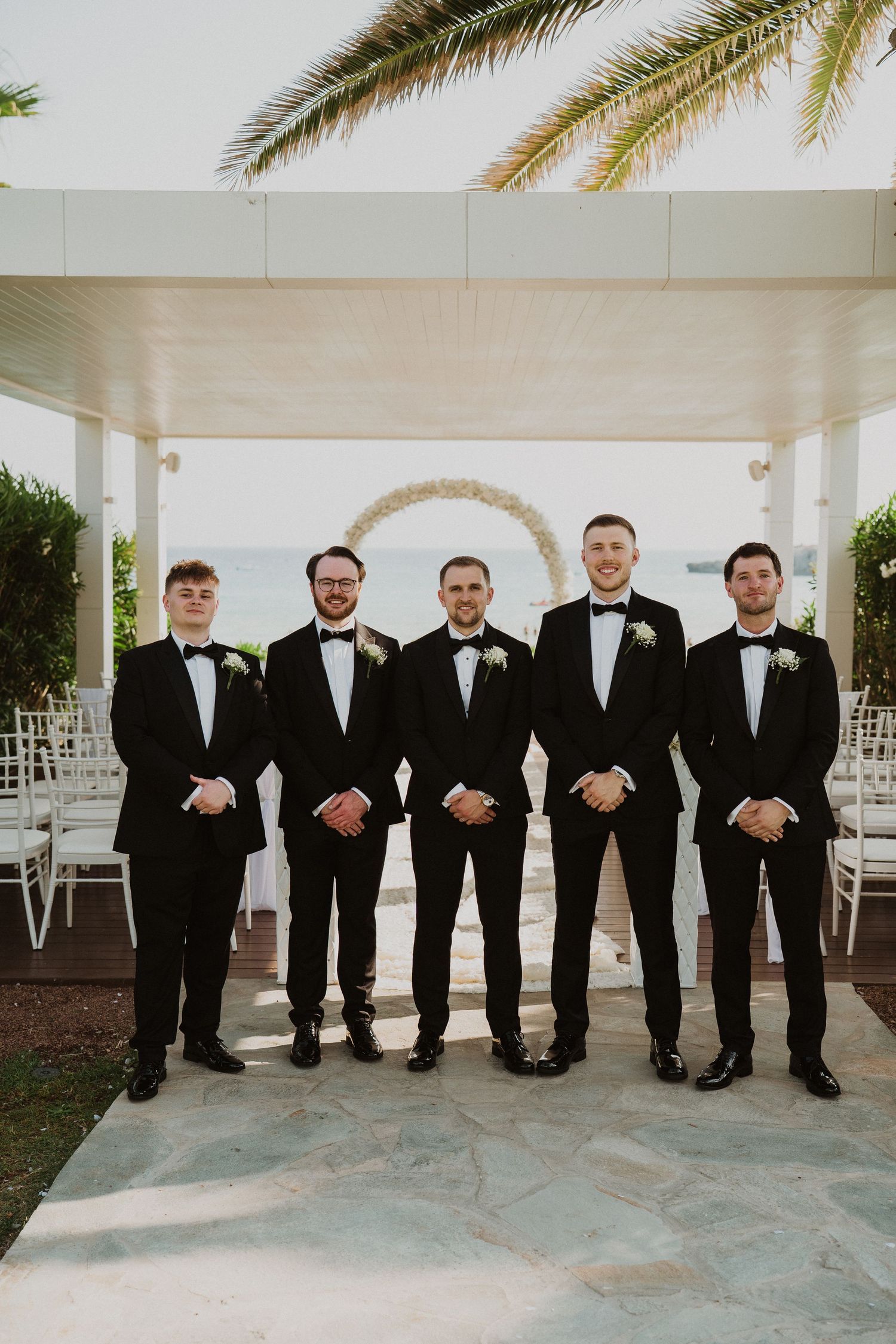 Groom and four groomsmen in matching black tuxedos pose under pergola with circular floral arch and ocean backdrop.