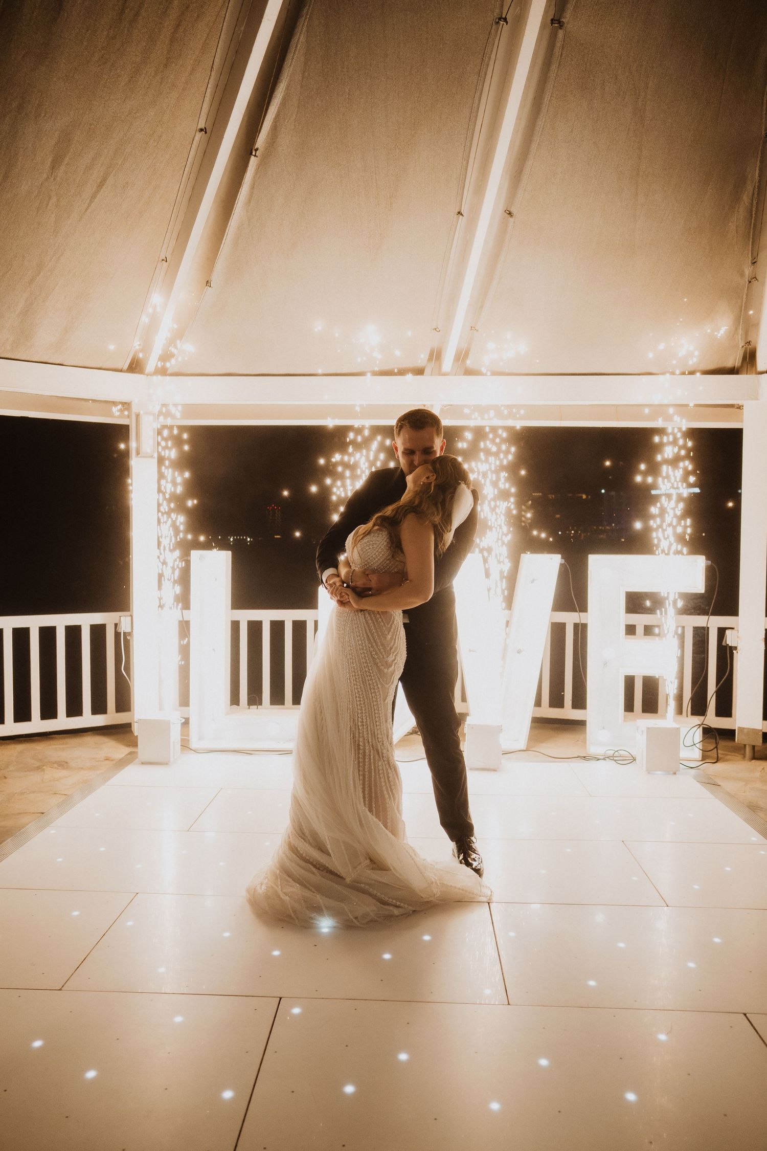 Bride and groom share first dance under warm fairy lights with glowing LOVE letters behind them.