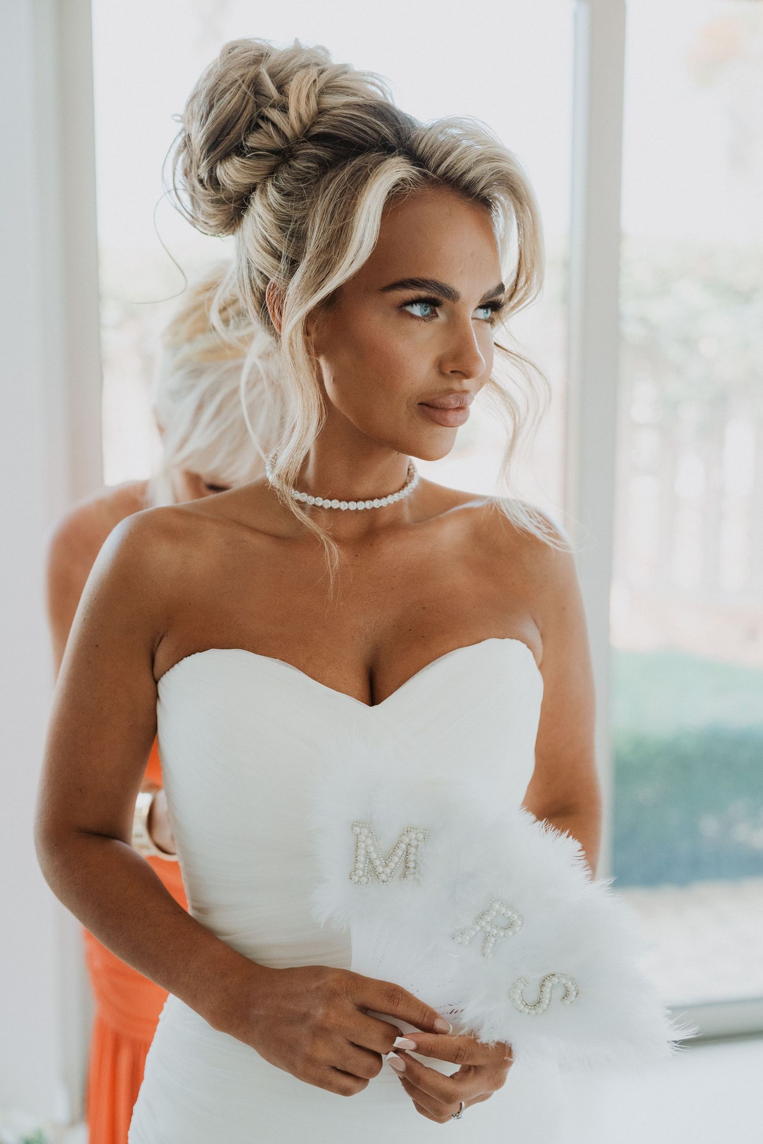 Bride in strapless white gown holding MRS feather fan, pearl choker necklace, elegant updo hairstyle, wedding day portrait.